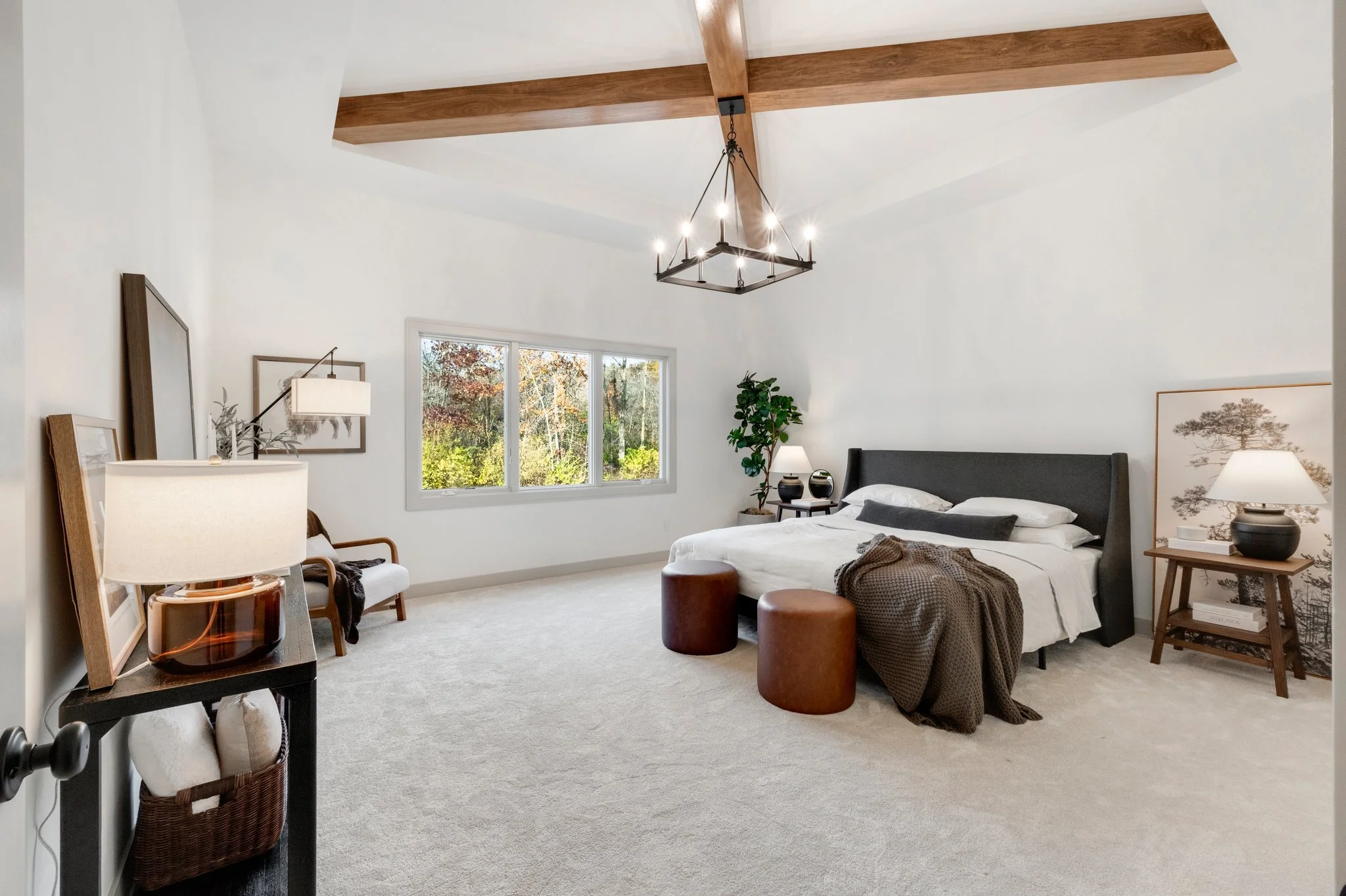 Modern bedroom with white walls, large window showing outdoor trees, black bed with white bedding, brown leather stools, and wooden ceiling beams.