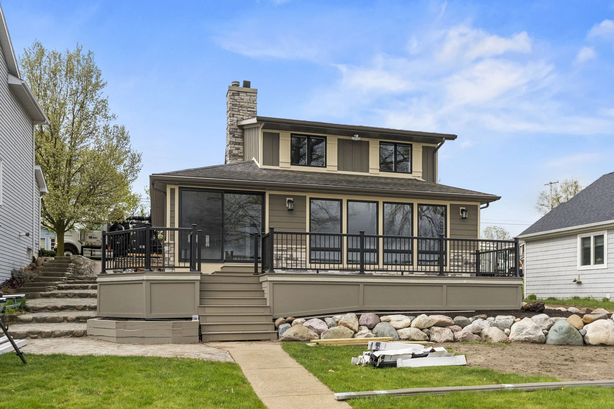 The image shows a two-story house with a deck and a set of stairs leading to the backyard. The house has large windows, a stone chimney, and siding that is beige with accents. There are neighboring houses on either side, green grass, a tree, and construction materials in the yard indicating ongoing work.