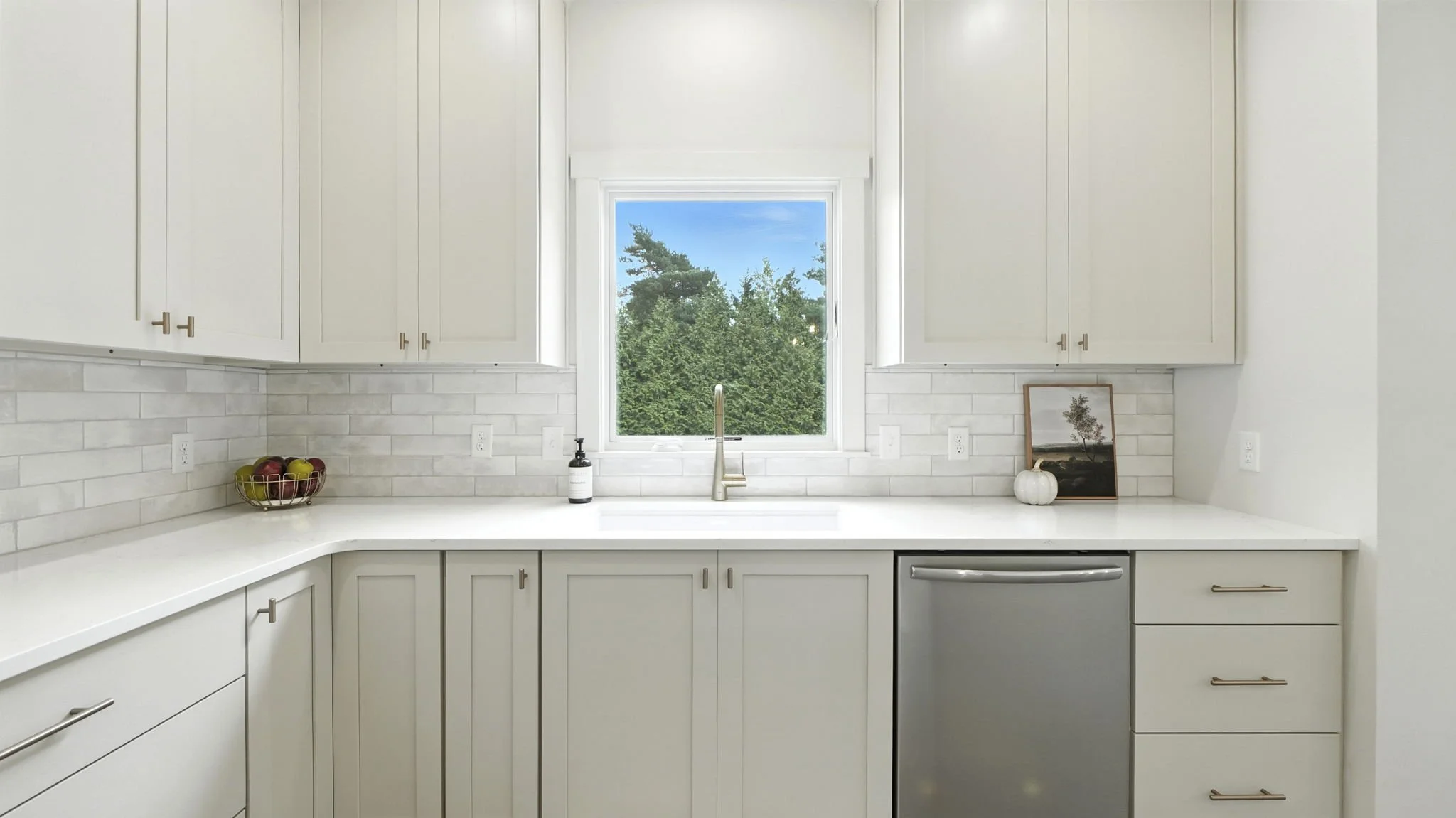 Bright kitchen with white cabinets, a window overlooking trees, and a marble backsplash. Countertop has a fruit bowl, soap dispenser, framed picture, and pumpkin decor. Stainless steel dishwasher below the counter.