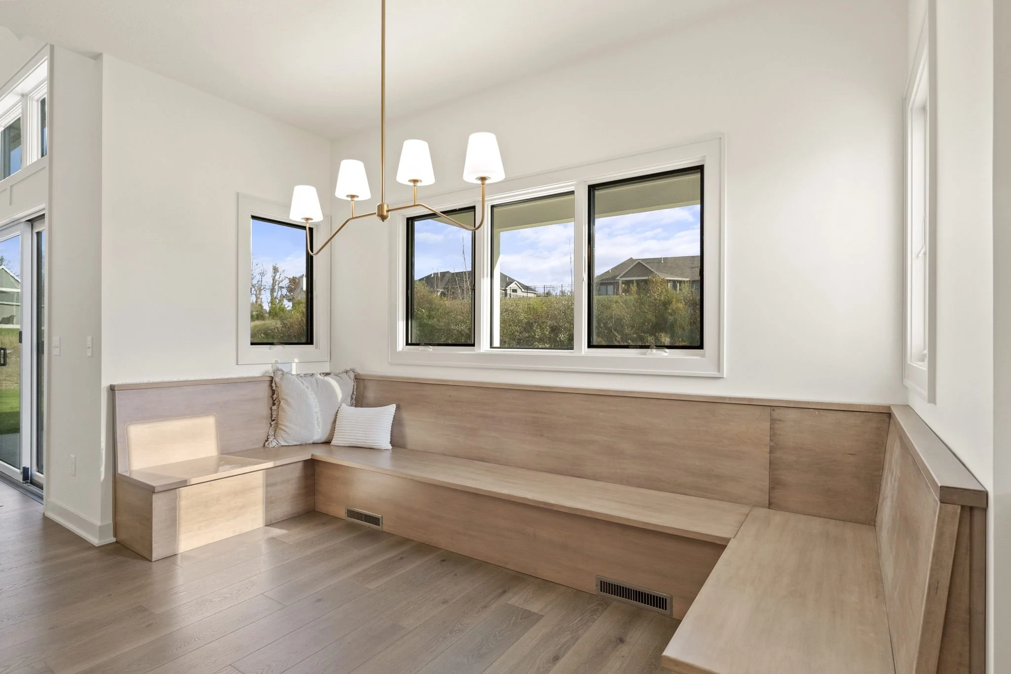 Modern living room with built-in light wood seating along the walls, three large windows showing a suburban neighborhood, white walls, a white ceiling, hardwood floors, and a modern chandelier with six lamps.