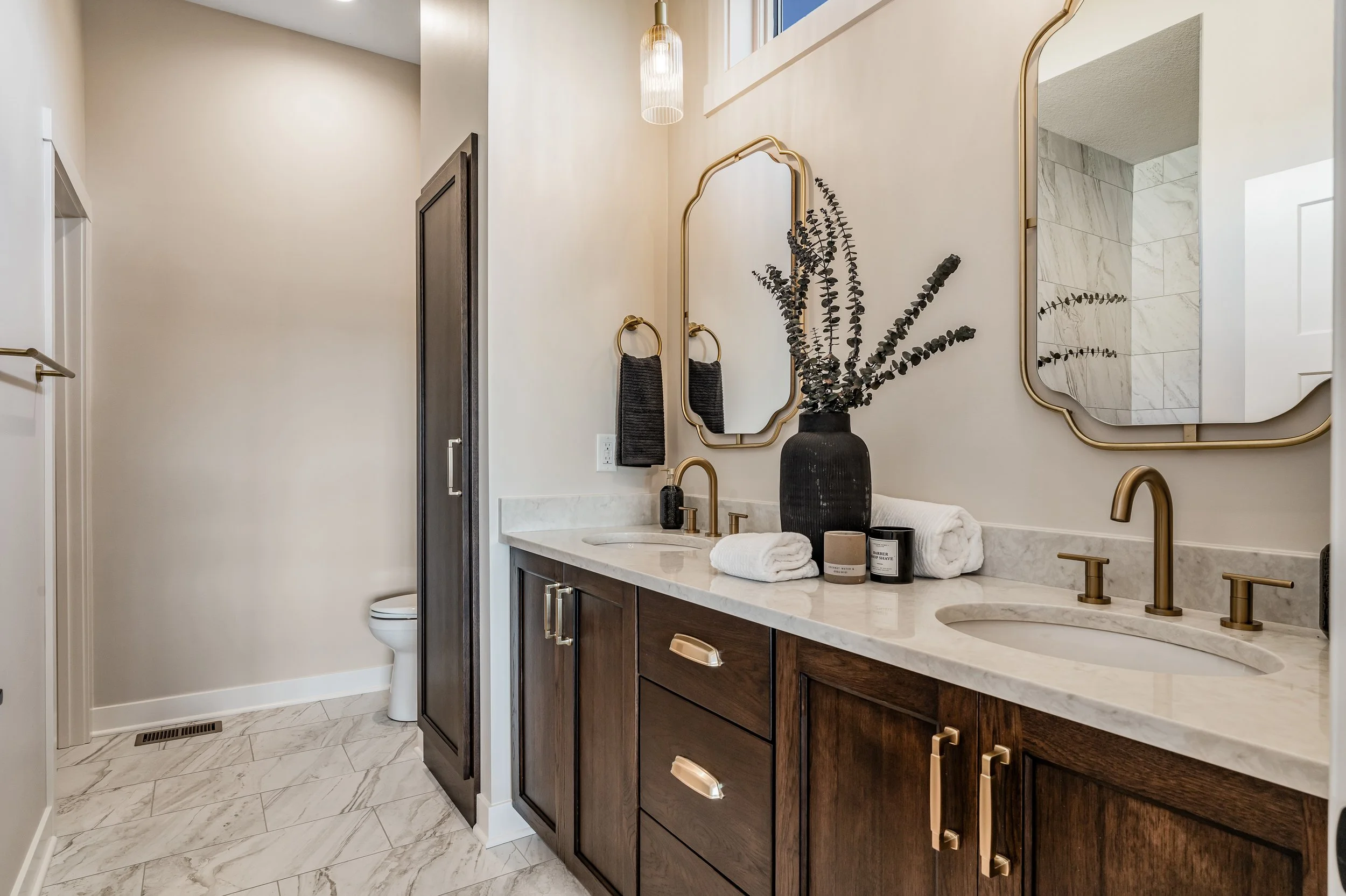 Modern bathroom with a double vanity, dark wood cabinets, white marble countertop, two oval mirrors with gold frames, black vase with greenery, rolled towels, candles, and black and gold fixtures.