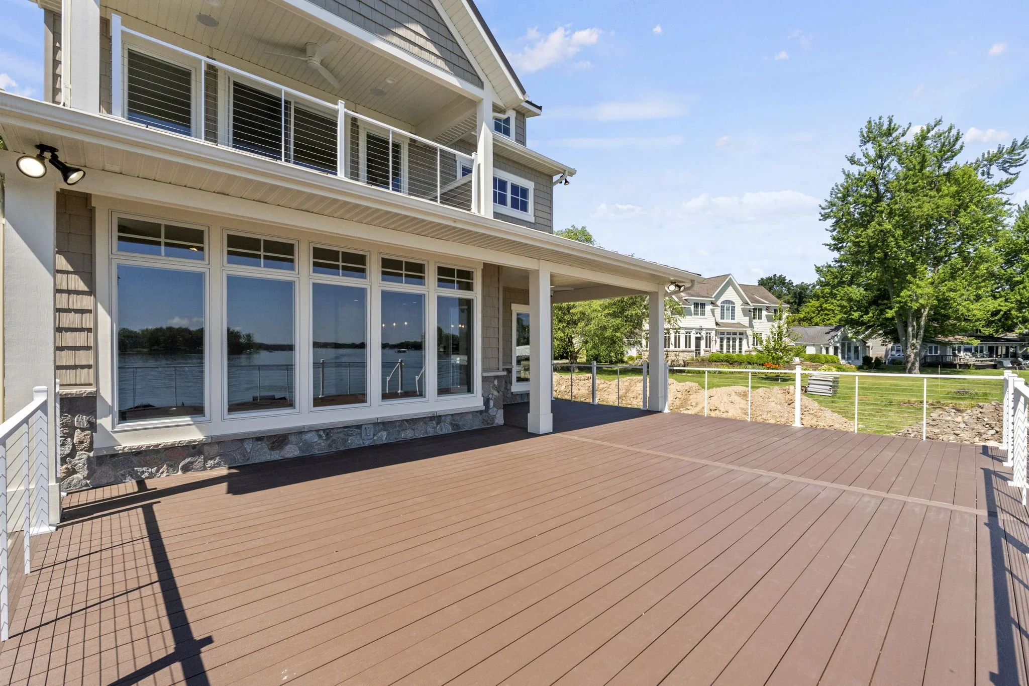 Large wooden deck outside a house featuring large windows, a covered porch area, and a view of neighboring houses across a grassy yard with trees.