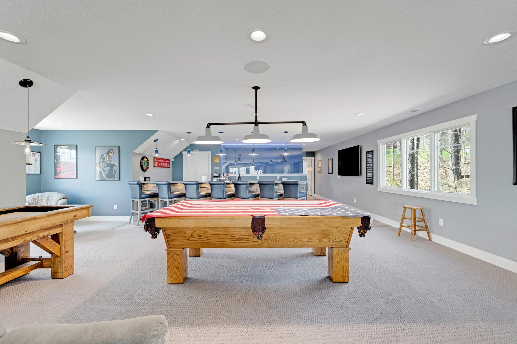 A spacious basement entertainment room with a pool table draped with an American flag, a bar with barstools, wall-mounted TV, framed posters, a dartboard, and large windows showing a wooded outdoor area.