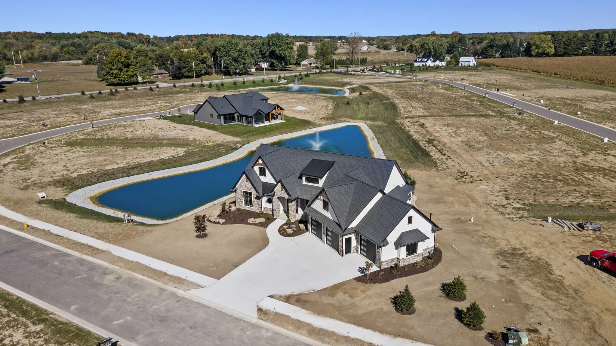 Aerial view of a modern house with a large driveway, adjacent to a pond with a fountain, and additional ponds and houses in the background in a rural setting.