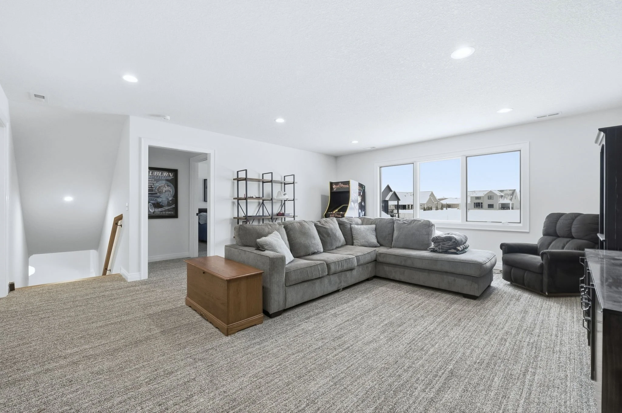Living room with a large beige sectional sofa, a small wooden coffee table, gray and black armchairs, a wall with windows showing houses outside, a vintage arcade game, and a black bookshelf with decorative items.