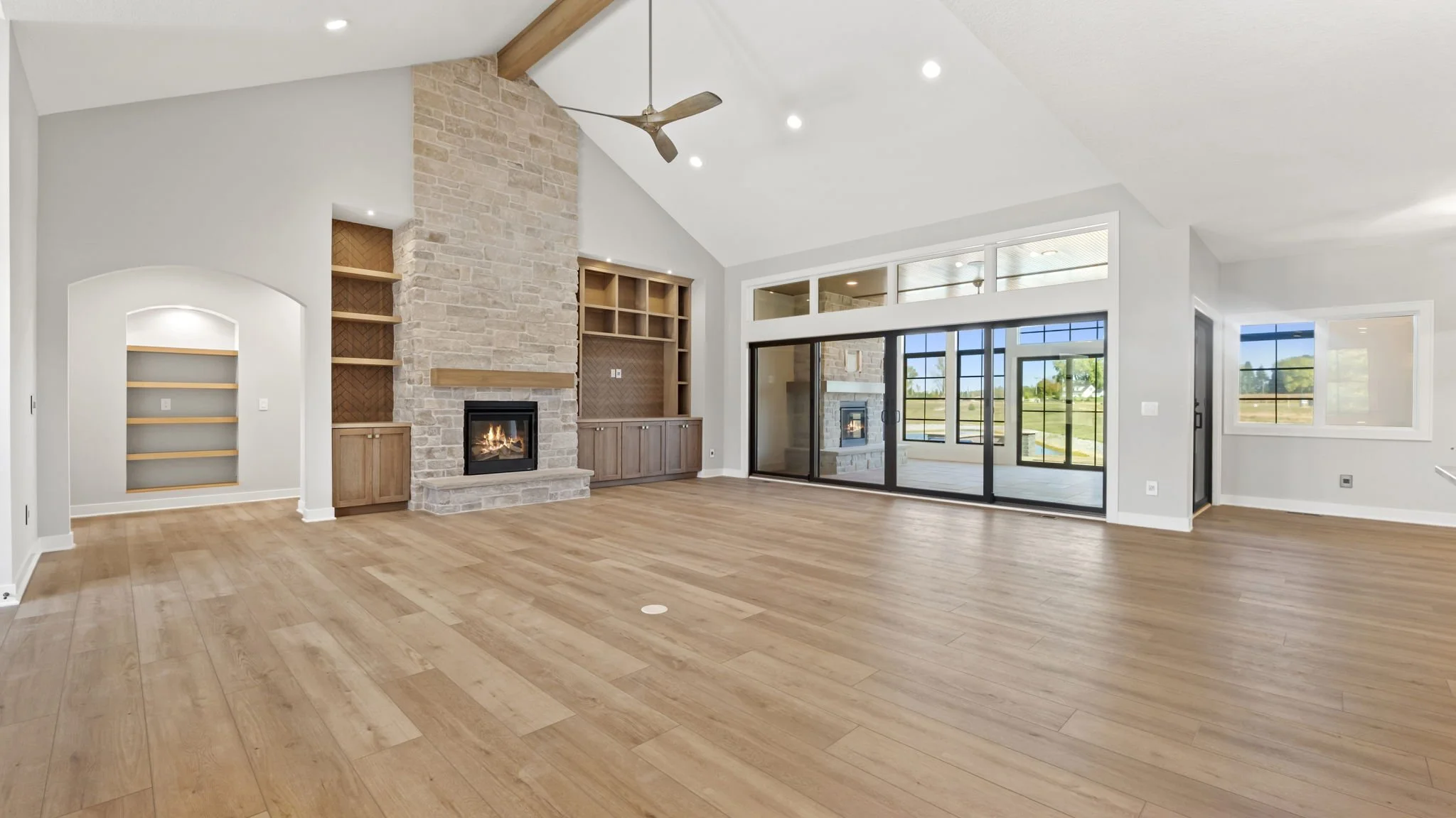 Spacious living room with a vaulted ceiling, wood flooring, a large stone fireplace, built-in wooden shelves, and sliding glass doors leading to an outdoor patio, with panoramic views of a grassy landscape.