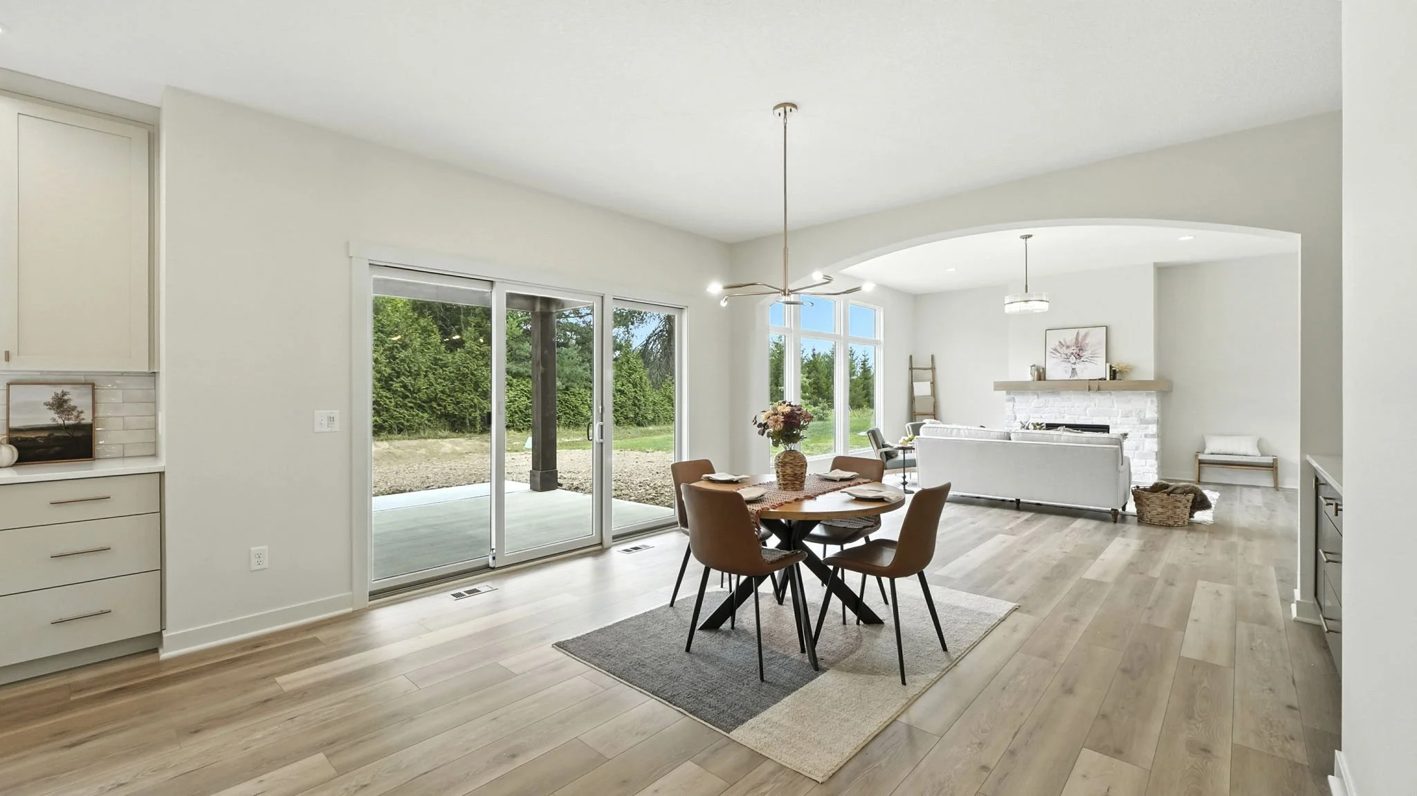 Open living and dining area with light wood flooring, white walls, sliding glass door leading outside, and a white brick fireplace with a mantle in the background.