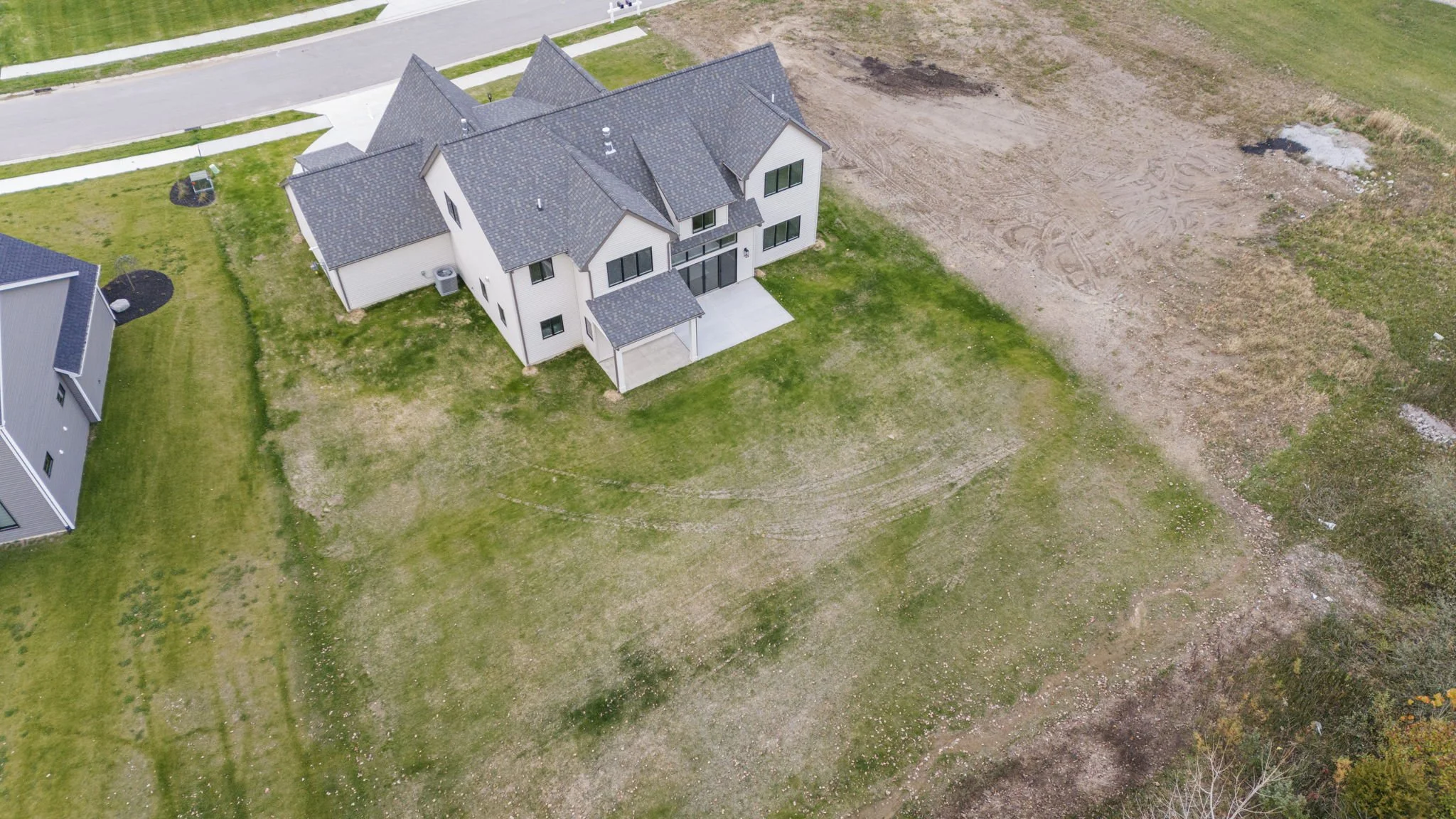 Aerial view of a modern two-story house with a dark gray roof and white exterior walls, surrounded by a spacious grassy yard with some patches of dirt and unfinished landscaping.