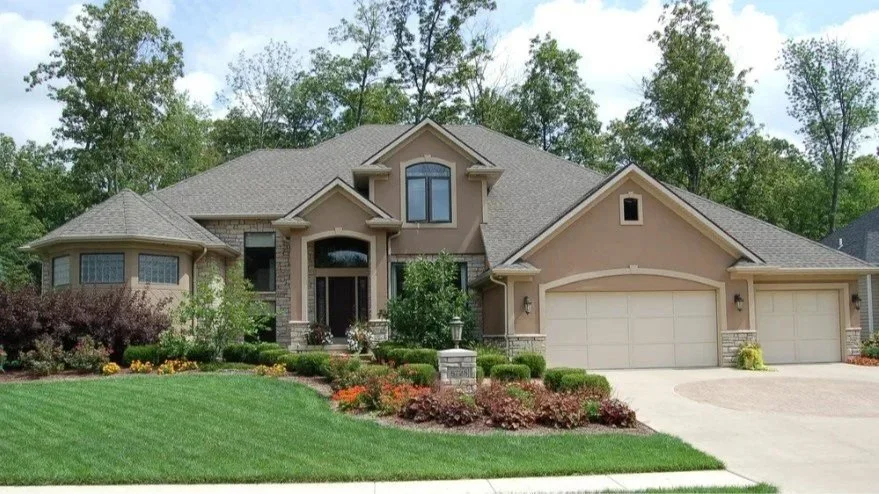 A large, two-story suburban house with a manicured lawn and landscaped front yard, featuring a two-car garage and multiple windows.