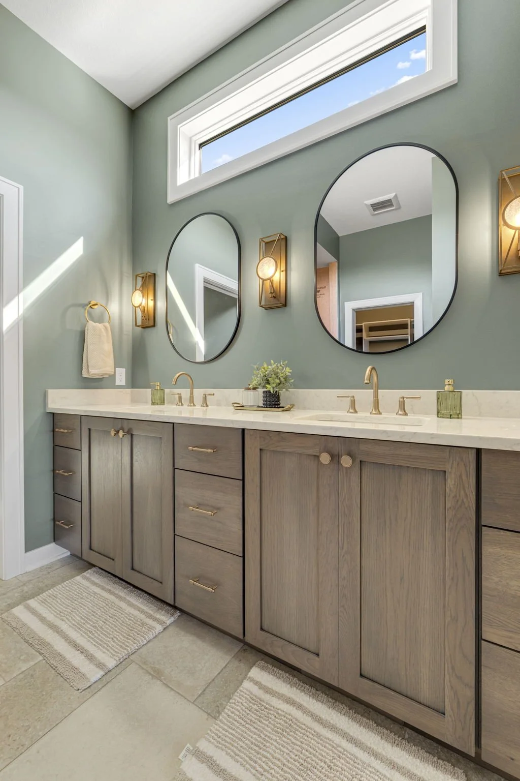 Bathroom with double vanity, gold fixtures, oval mirrors, wall-mounted light fixtures, green walls, a high window, and beige rugs.