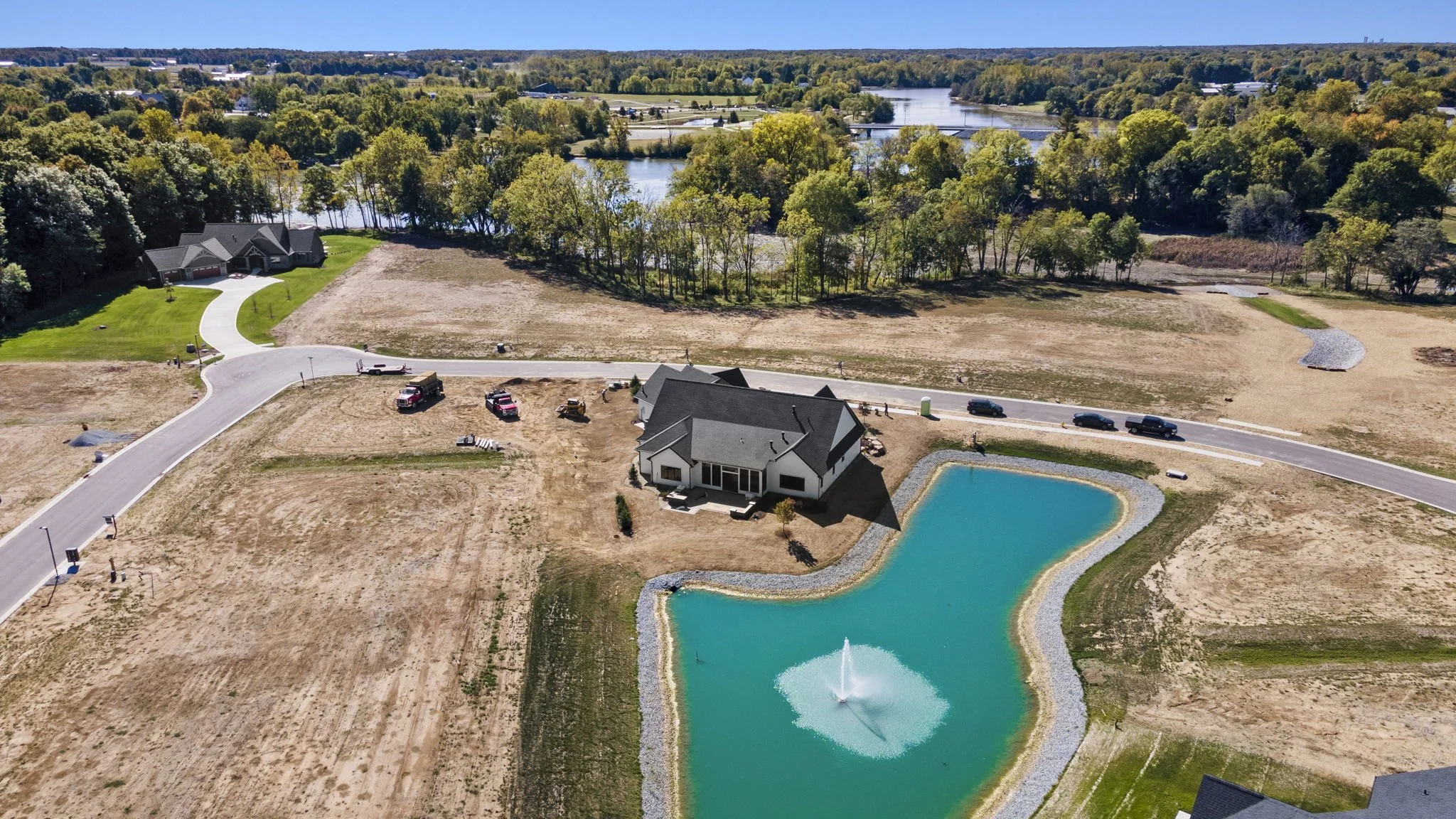 Aerial view of a modern house with a backyard pond featuring a fountain, surrounded by an open landscape with paved roads, sparse vegetation, and a nearby lake.
