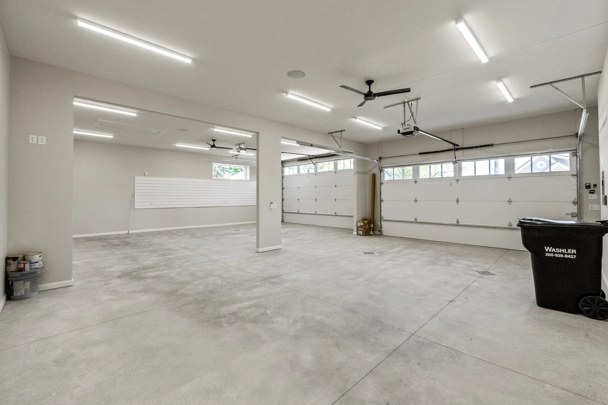 Empty spacious garage with two white garage doors, concrete floor, drywall walls, ceiling fans, and bright fluorescent lighting.