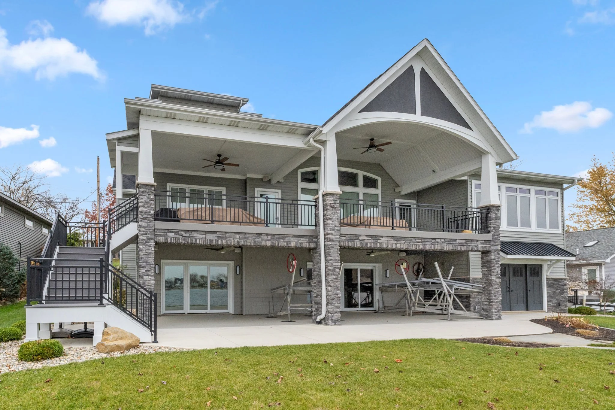 Two-story house with a large front patio, grey siding, stone accents, black metal railings, and multiple windows. There are stairs leading up to the upper balcony on both sides, with outdoor furniture and ceiling fans visible. The yard is well-mainta