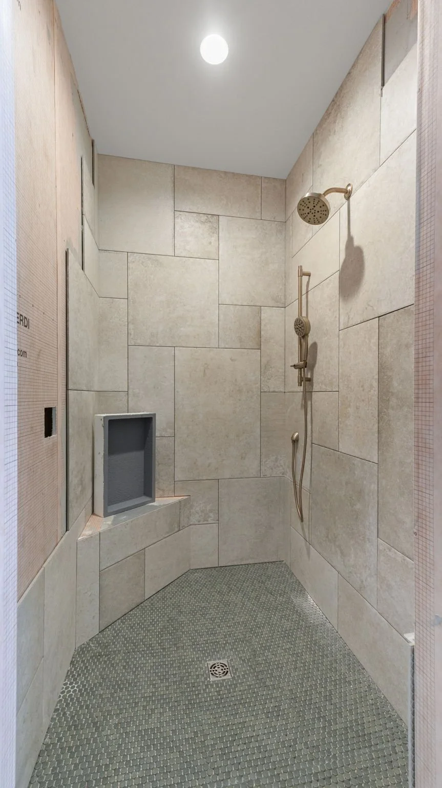 Empty shower with beige tiled walls, a single overhead light, a showerhead, a handheld shower wand, a small built-in shelf, and a metal drain cover on the textured floor.