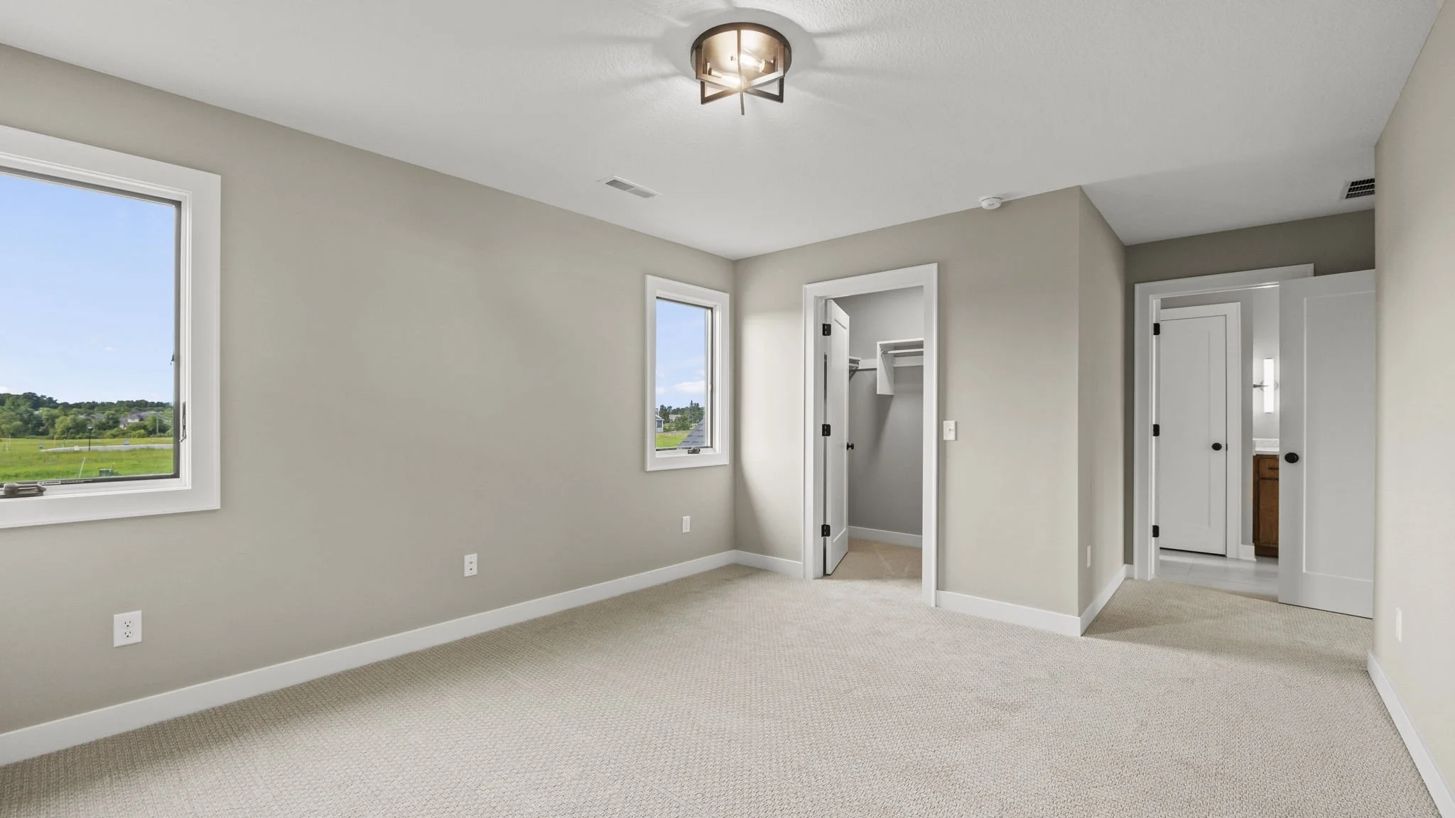 Empty bedroom with beige carpet, light-colored walls, two windows showing a green landscape, ceiling light fixture, and an open closet with shelves and a rod.