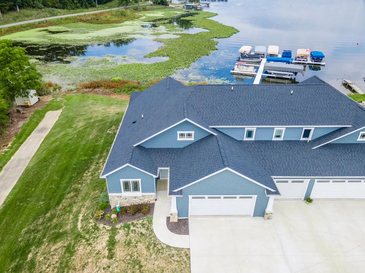 Aerial view of a blue house with a gray roof next to a marina with boats on the water.