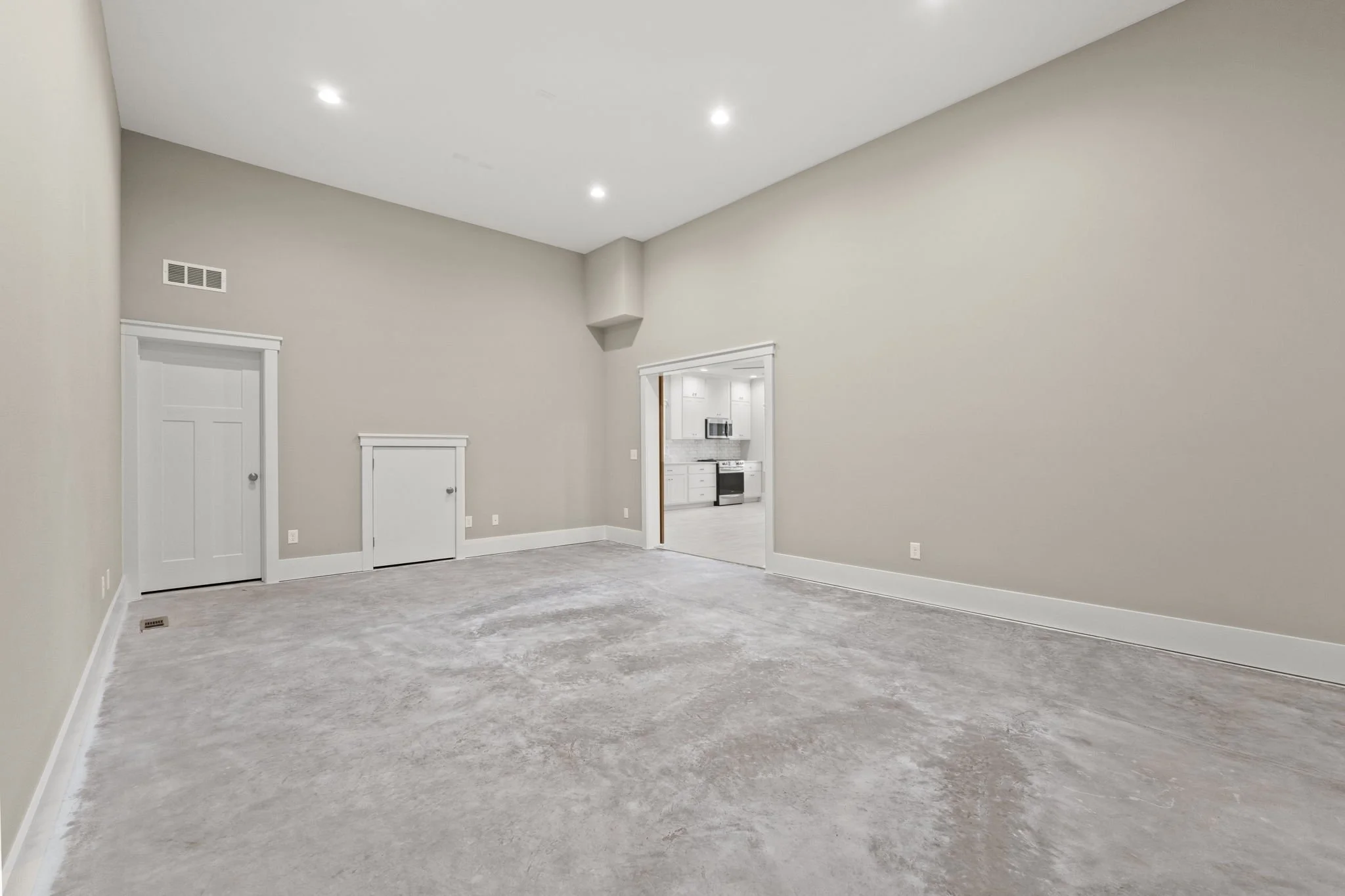 Empty living room with beige walls, light gray carpet, white baseboards, and ceiling, with a doorway leading to a kitchen with white cabinets and stainless steel appliances.