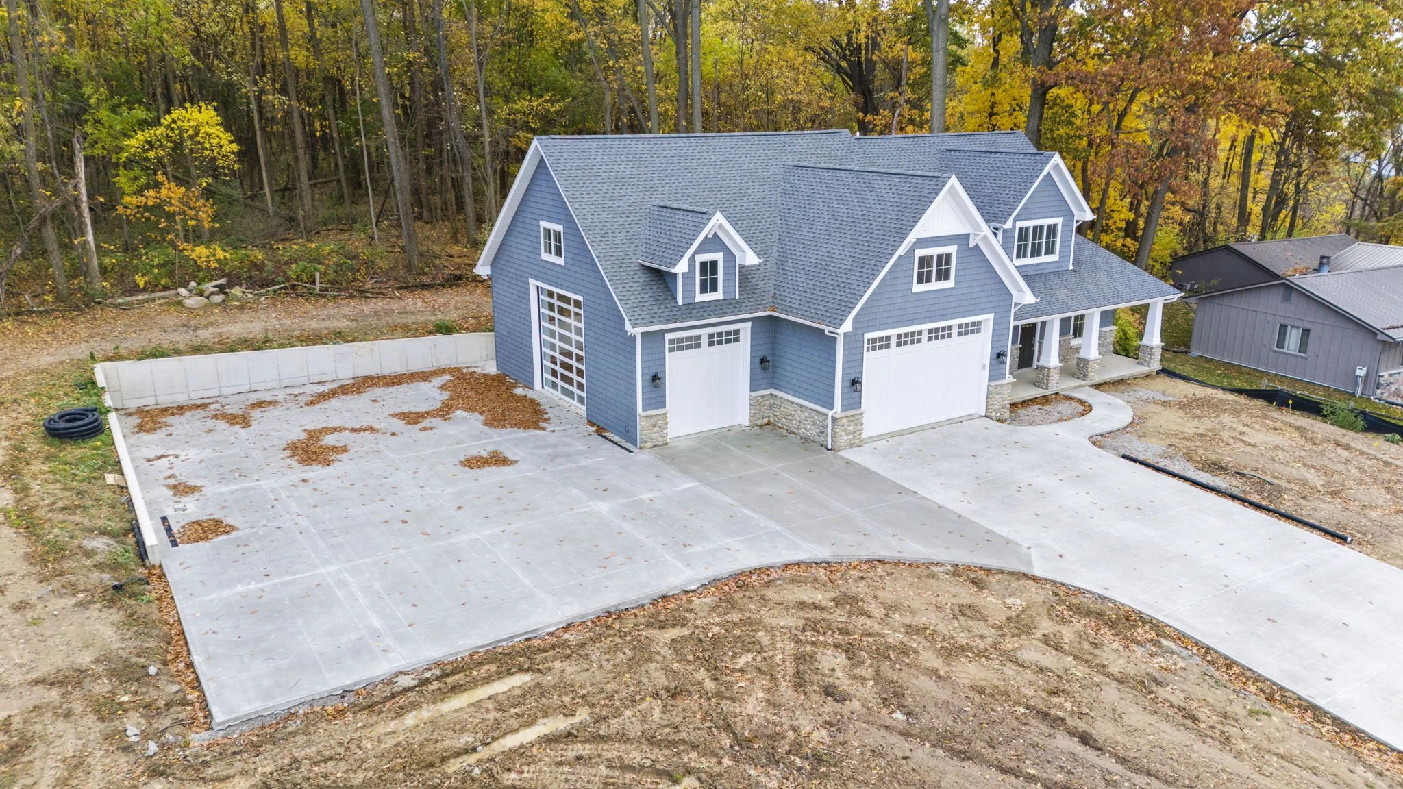 A newly built blue house with a front porch and attached garage, surrounded by a large concrete driveway, with a wooded area in the background.