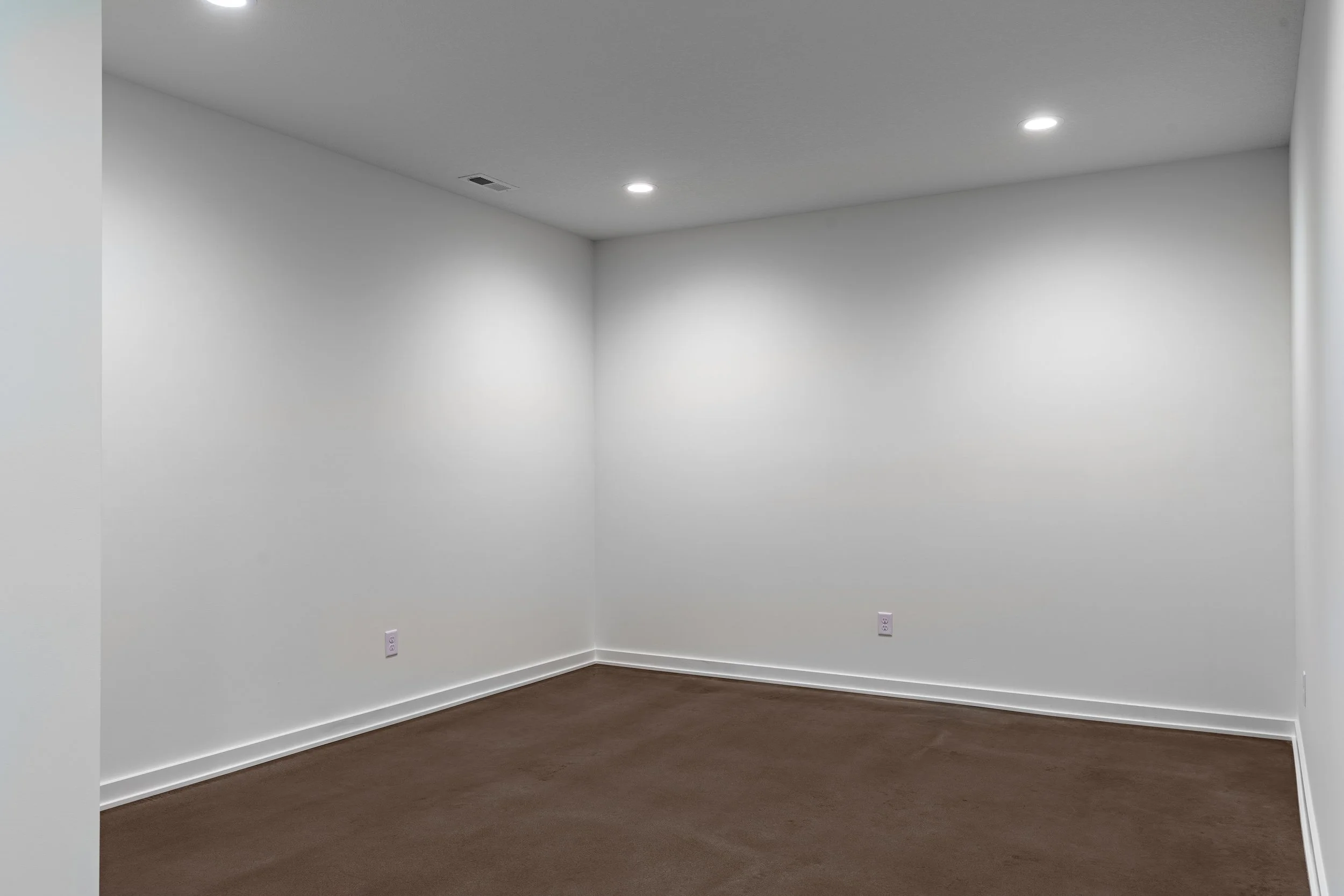 Empty room with white walls, brown carpet, and recessed ceiling lights.