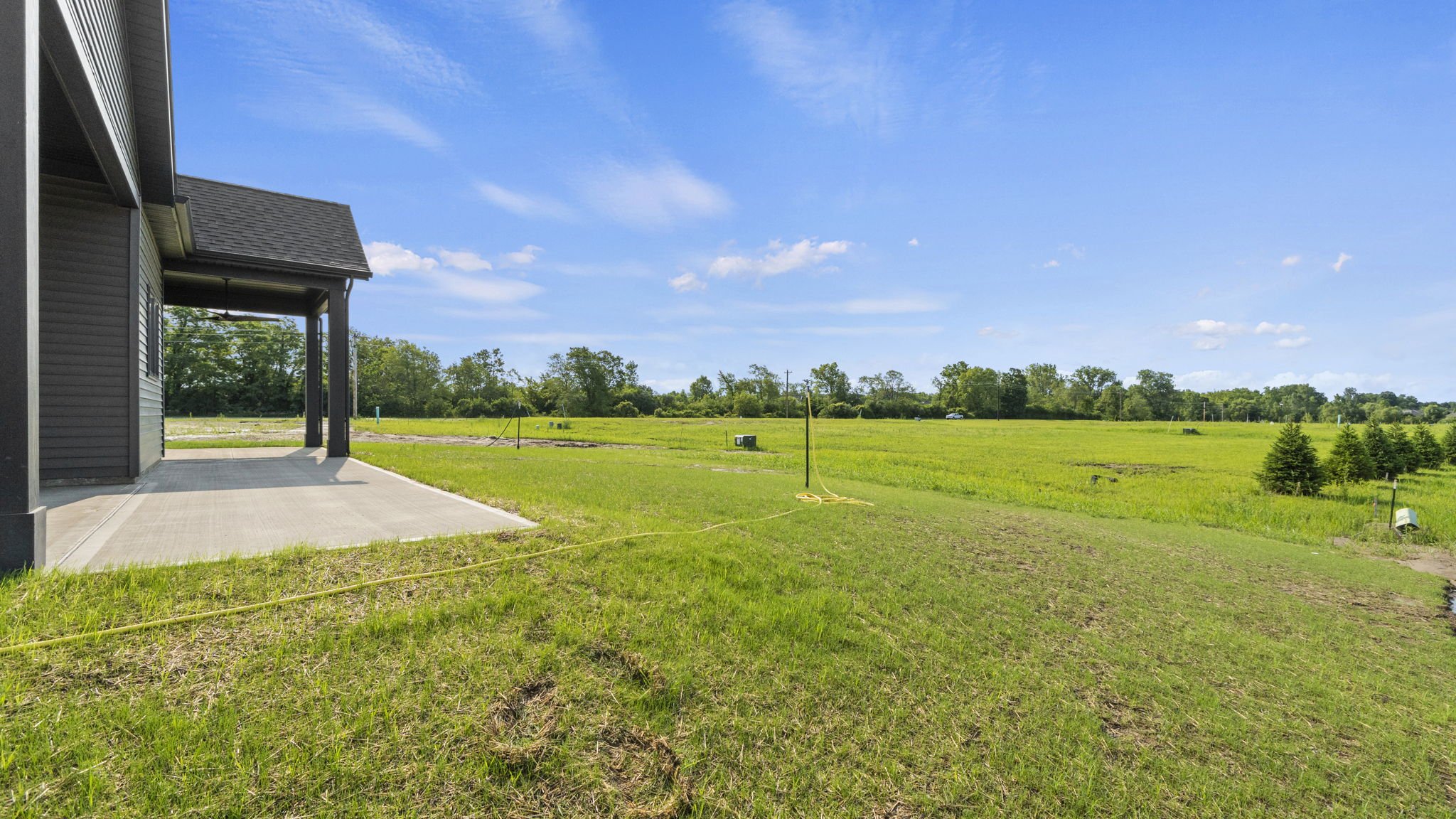 View of a backyard with a concrete patio attached to a house on the left, and a large open grassy field with some small trees and a blue sky with white clouds in the distance.