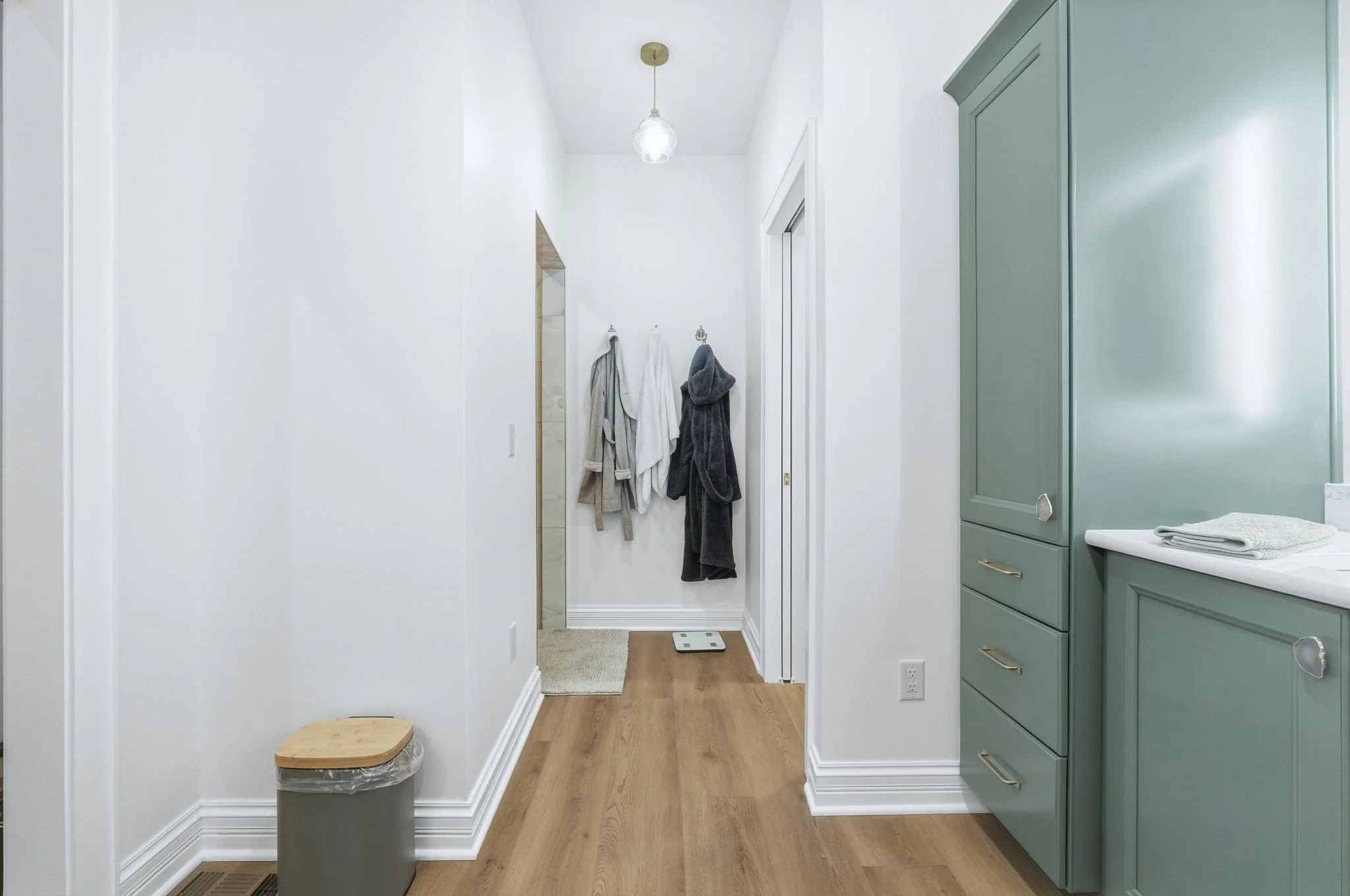 A hallway leading to a door with towels hanging on hooks, a small step stool, a wall-mounted thermostat, a digital bathroom scale, and a green cabinet with drawers and a towel on top.
