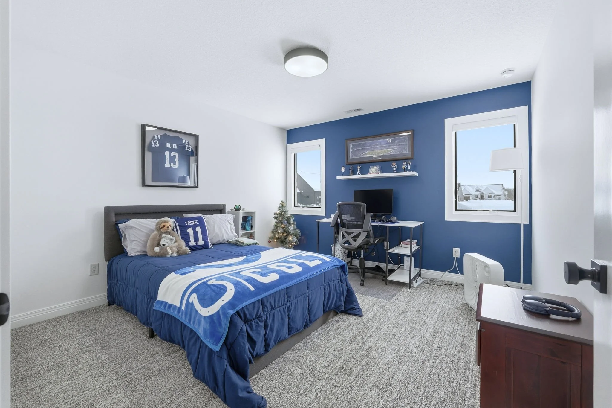A bedroom with a gray bed, blue and white bedding, and stuffed animals, the wall behind the bed is white, and the other wall is blue. There is a monitor, desk, and chair against the blue wall, with windows showing a snowy suburban neighborhood outsid