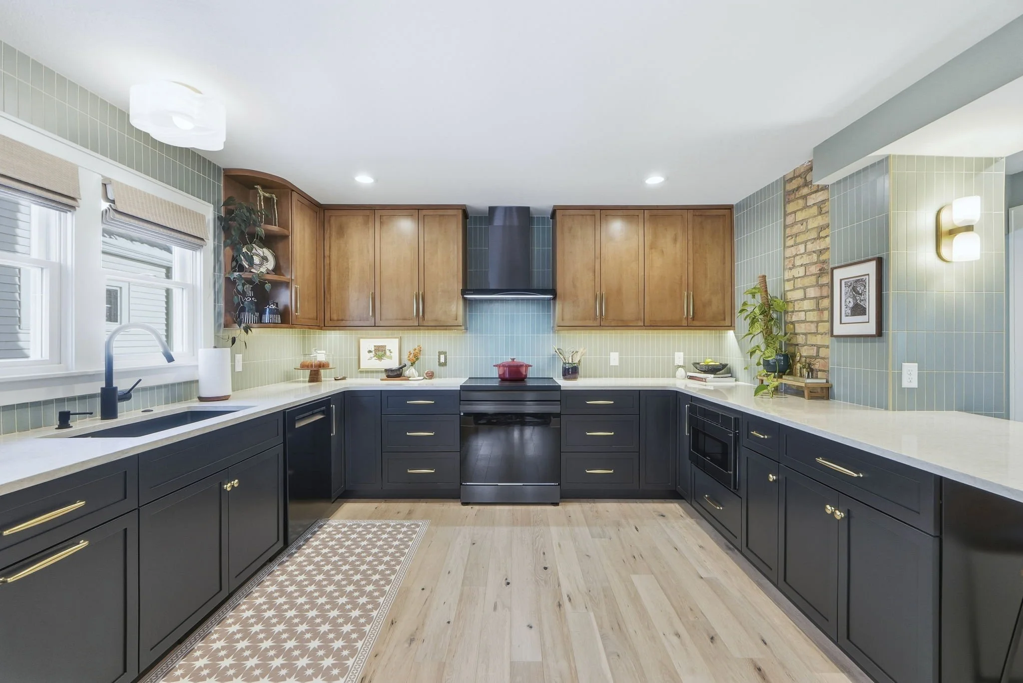 Modern kitchen with dark lower cabinets, wooden upper cabinets, a black stove in the center, and light-colored countertops. Features include a window with blinds, plants, framed art, and a brick accent wall.