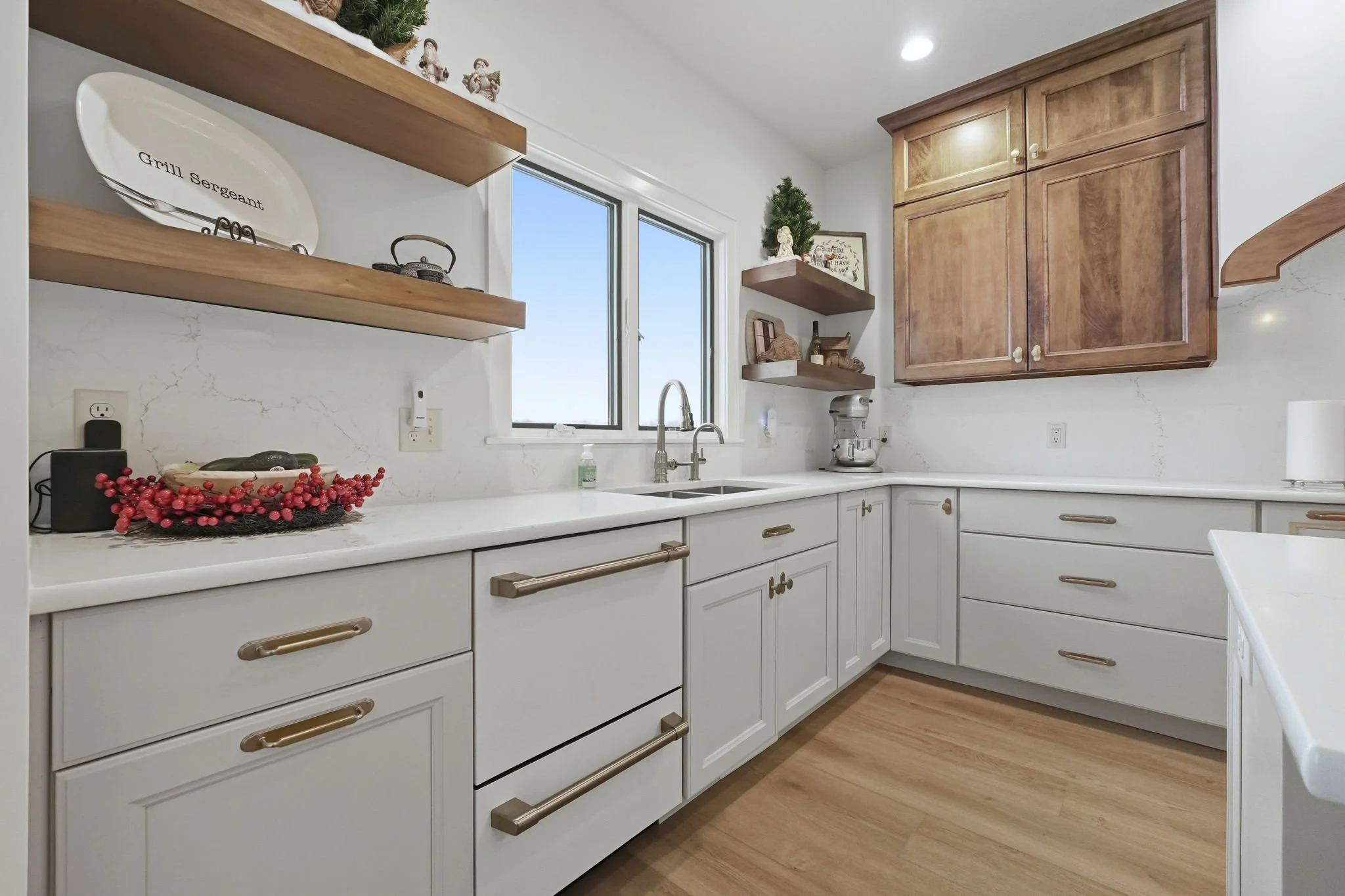 A kitchen with white cabinets, light wood flooring, and wooden open shelves. The shelves hold decorative items and a cooking item. There is a window above the sink, and a countertop with Christmas decorations and a stand mixer.