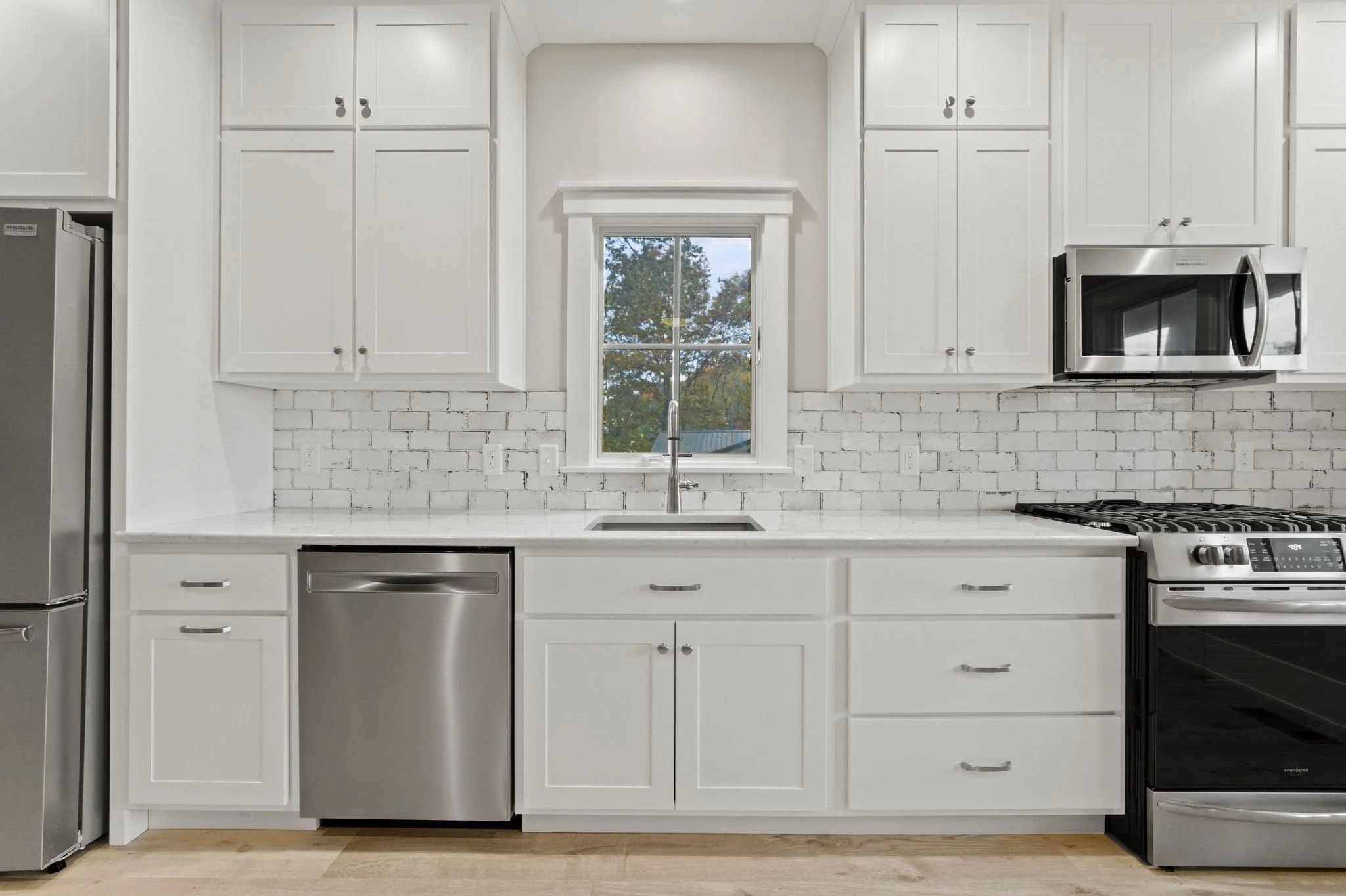 Modern white kitchen with stainless steel appliances, a window above the sink, and a brick backsplash.