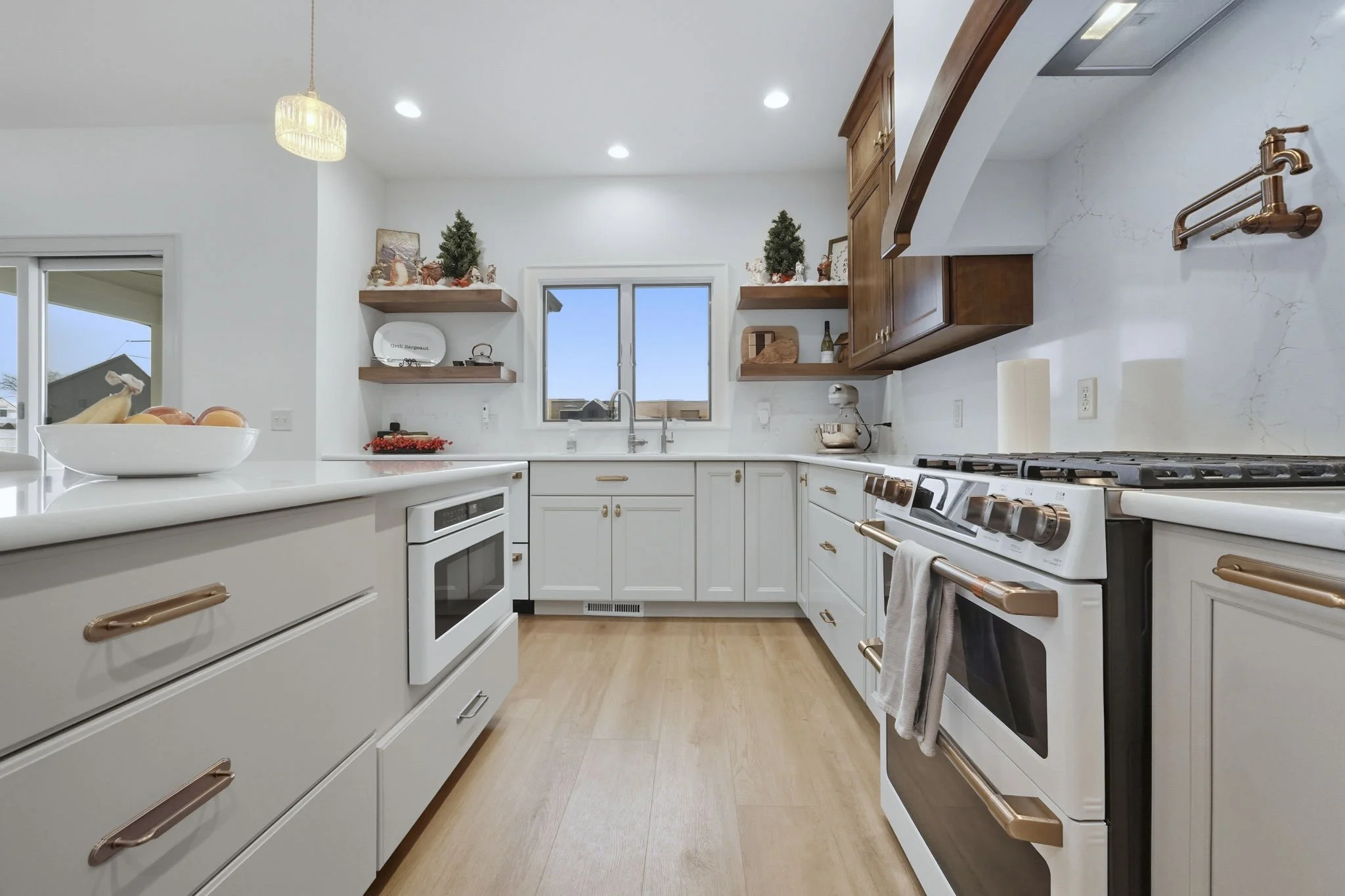Modern kitchen with white cabinets, wooden accents, and stainless steel appliances. Decor includes small potted trees and holiday decorations, with a window above the sink allowing natural light.
