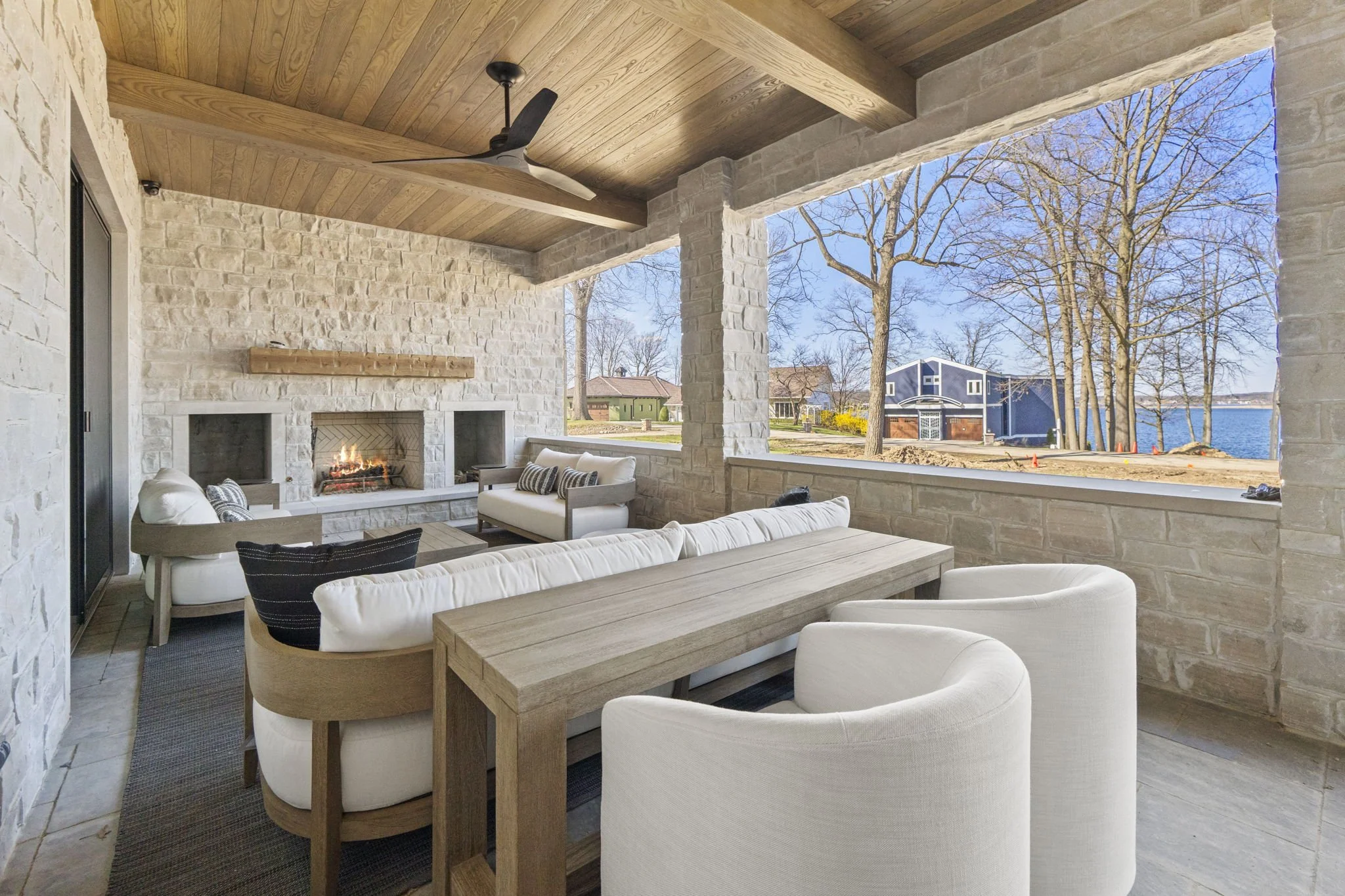 Outdoor covered patio with stone walls, wooden ceiling, and ceiling fan. Patio has white and beige seating Including a wicker armchair, a sofa, and a dining table with chairs. There is a fireplace built into the stone wall, with a fire burning. Large