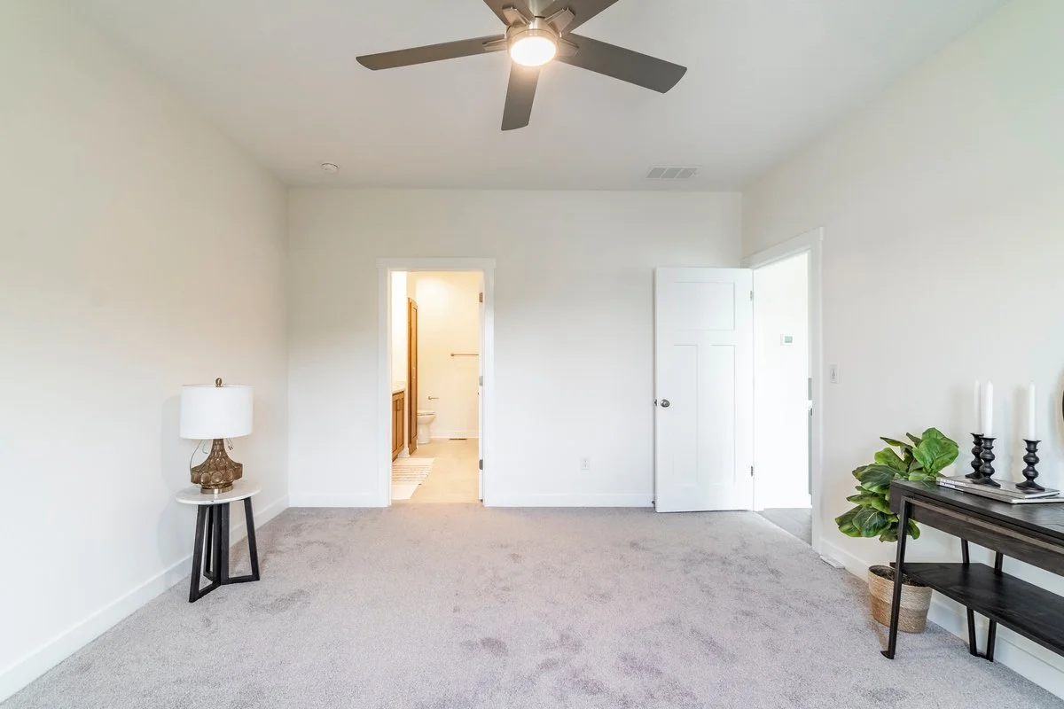 Empty living room with gray carpet, ceiling fan, white walls, and a small side table with a lamp. An open doorway reveals a bathroom with a toilet and vanity, and another door leads to an exterior or closet. There is a black console table with candle