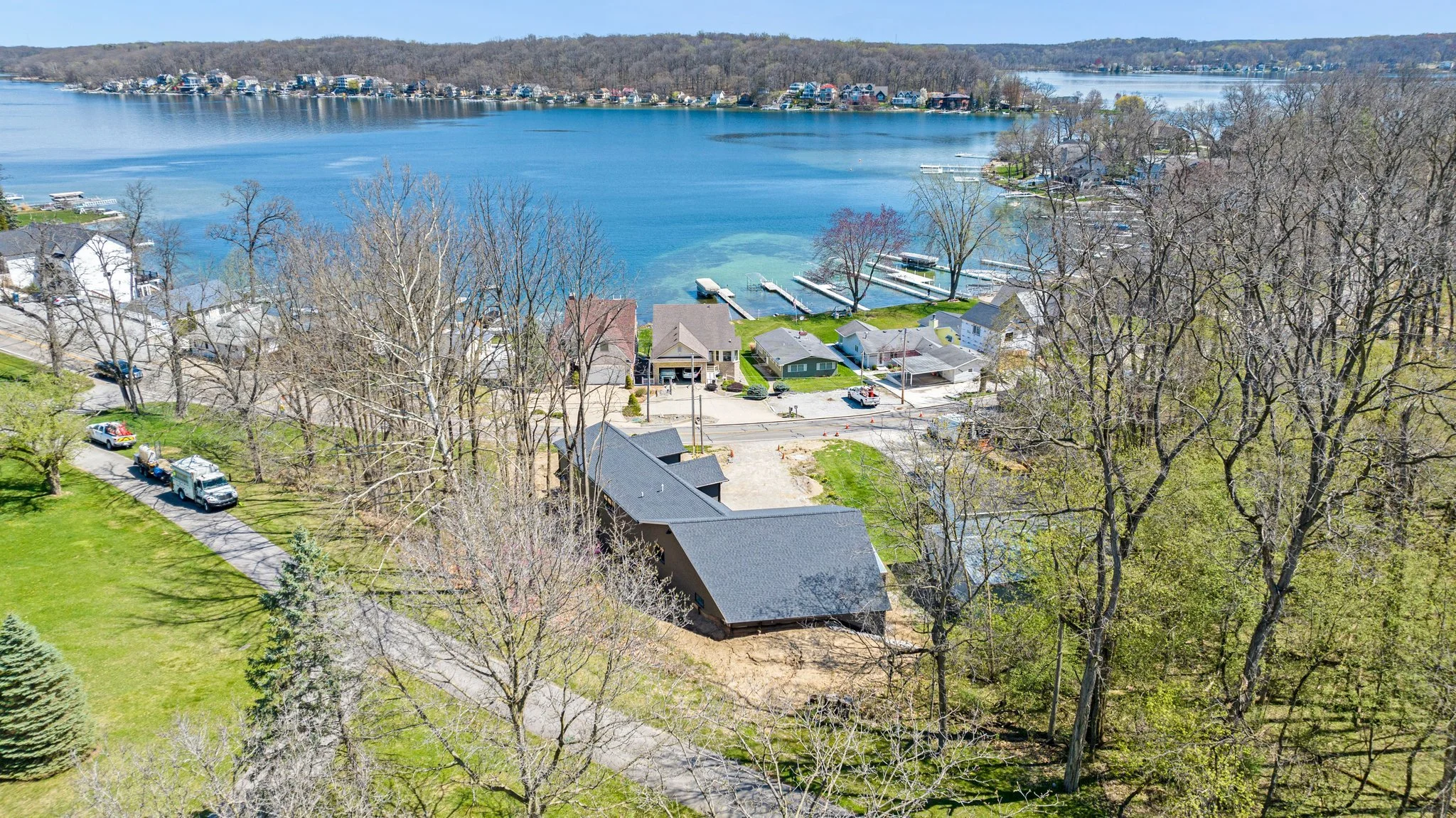 Aerial view of a lakeside residential area in spring, showing a newly constructed house, other completed homes, a grassy yard with trees, a lake with docks, and a wooded area.