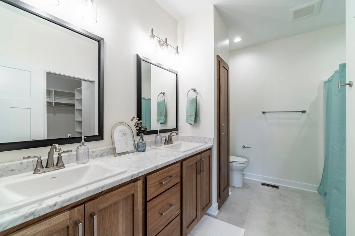 Bathroom with dual sinks, wooden cabinets, two mirrors, teal towels, and a toilet behind a wall, with a shower curtain on the right side.