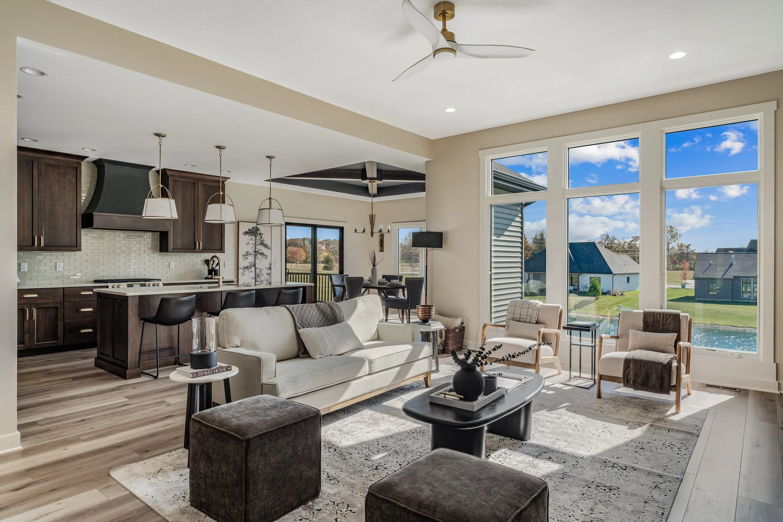 Open-concept living room and kitchen with large windows showing a backyard and blue sky, modern furniture, and pendant lighting.