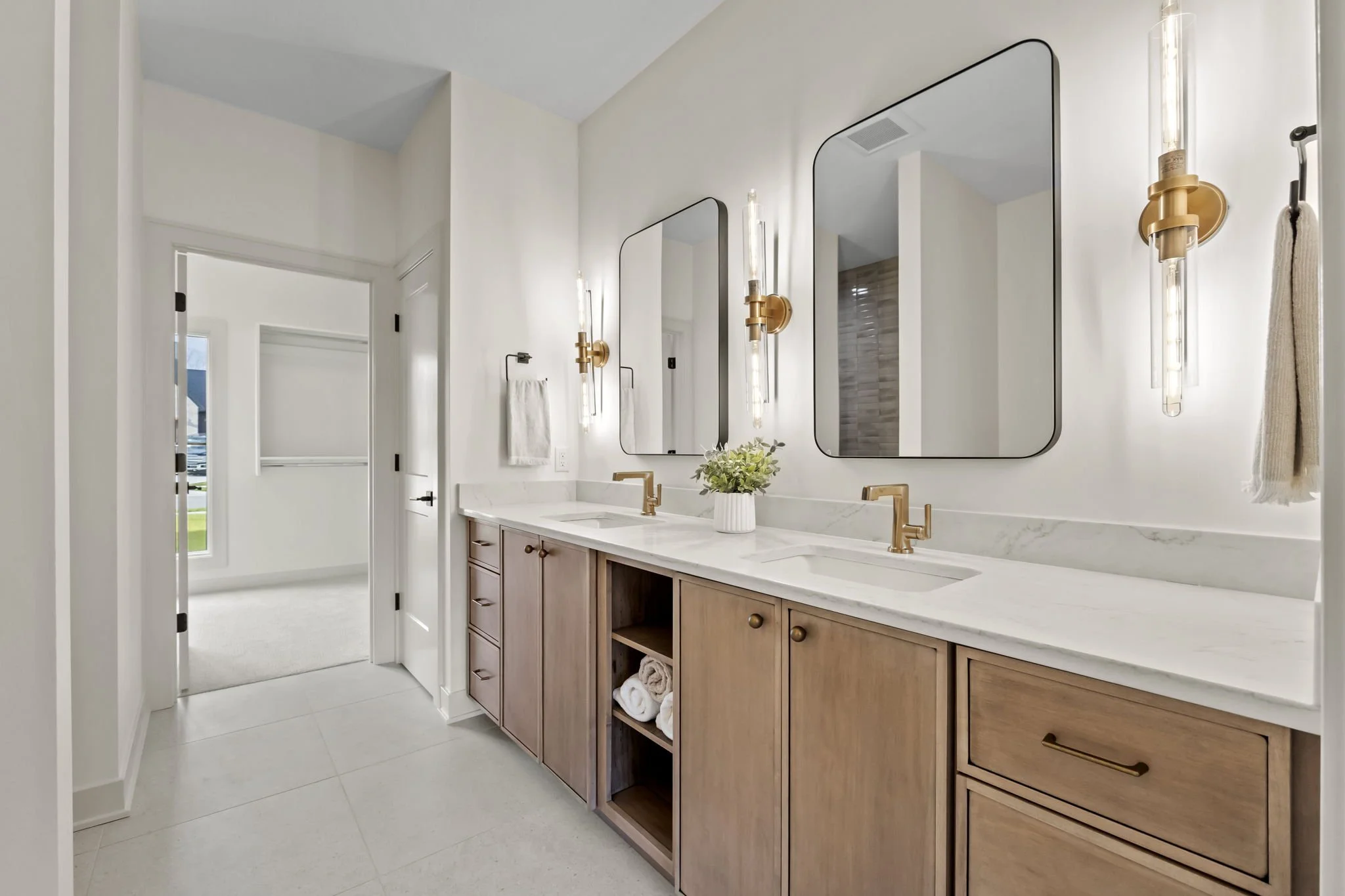 Modern bathroom with double vanity, large mirrors, brass fixtures, and wall-mounted light fixtures, with an adjacent room visible through the open door.