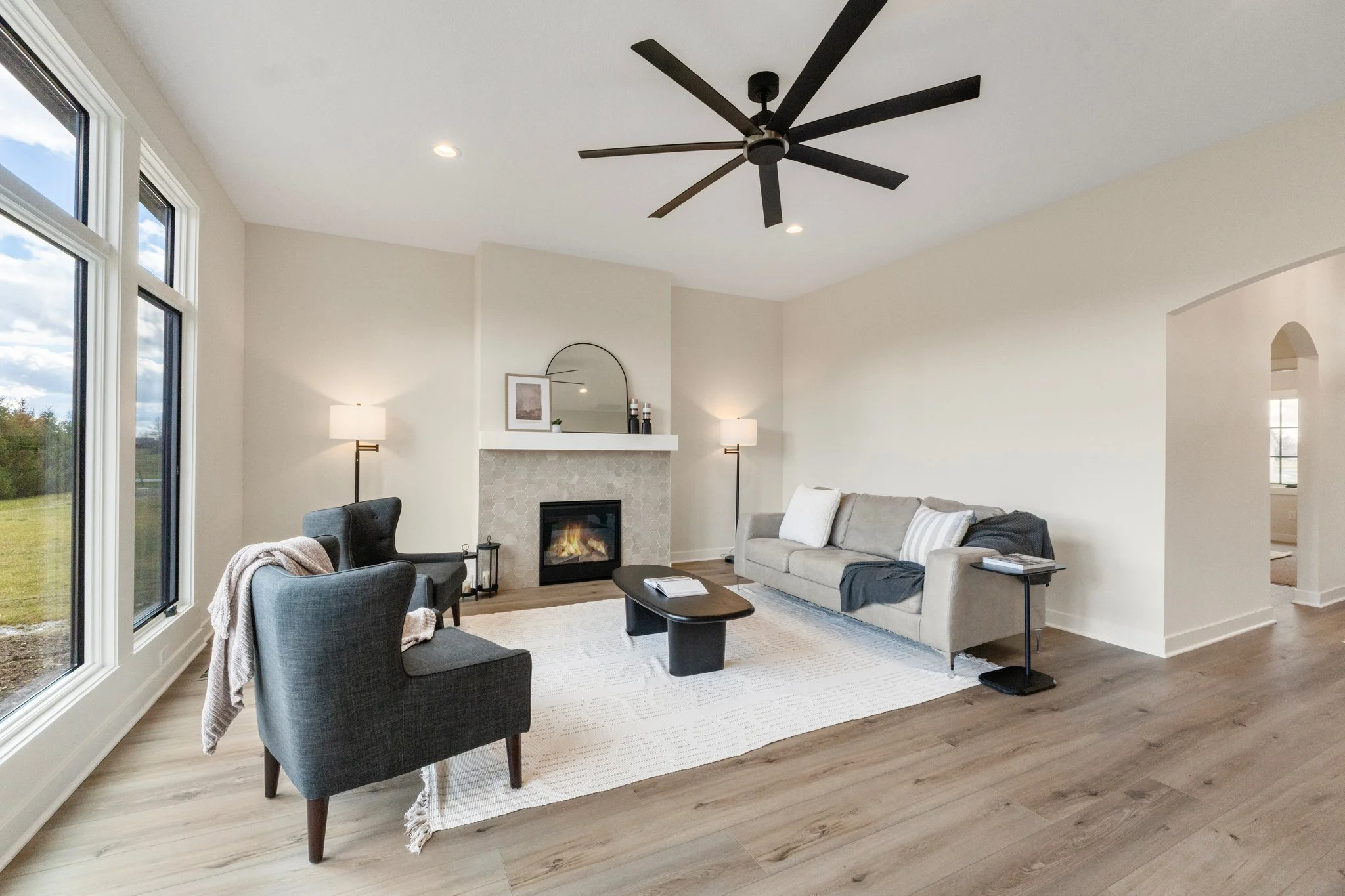 Modern living room with large windows, a fireplace, a gray sofa, two black armchairs, a white rug, and a black ceiling fan.