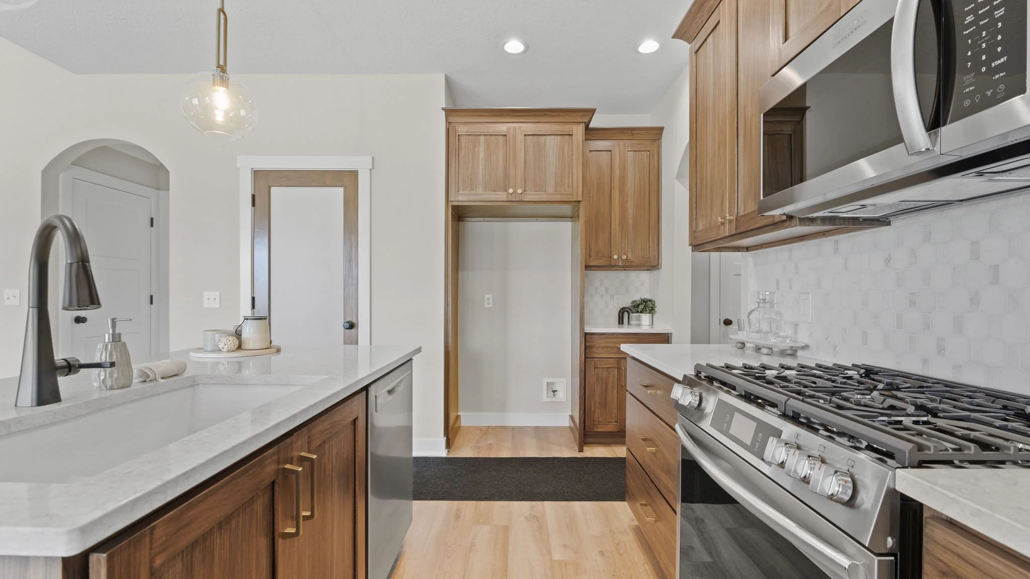 Modern kitchen with wooden cabinets, stainless steel appliances including a stove and microwave, white countertops, and a white tile backsplash. There is a double sink with a modern faucet, and some decorative items and small plants are on the counte