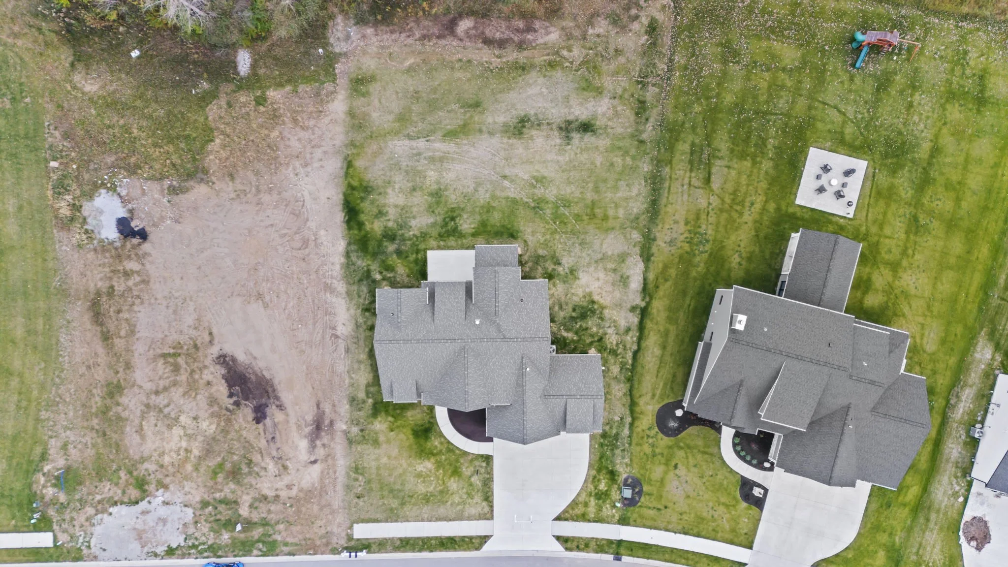 An aerial view of a residential backyard with two houses, one on the left and one on the right, separated by a grassy lawn. The left yard has patches of bare dirt and a small area with a blue object, while the right yard has a play structure with a s