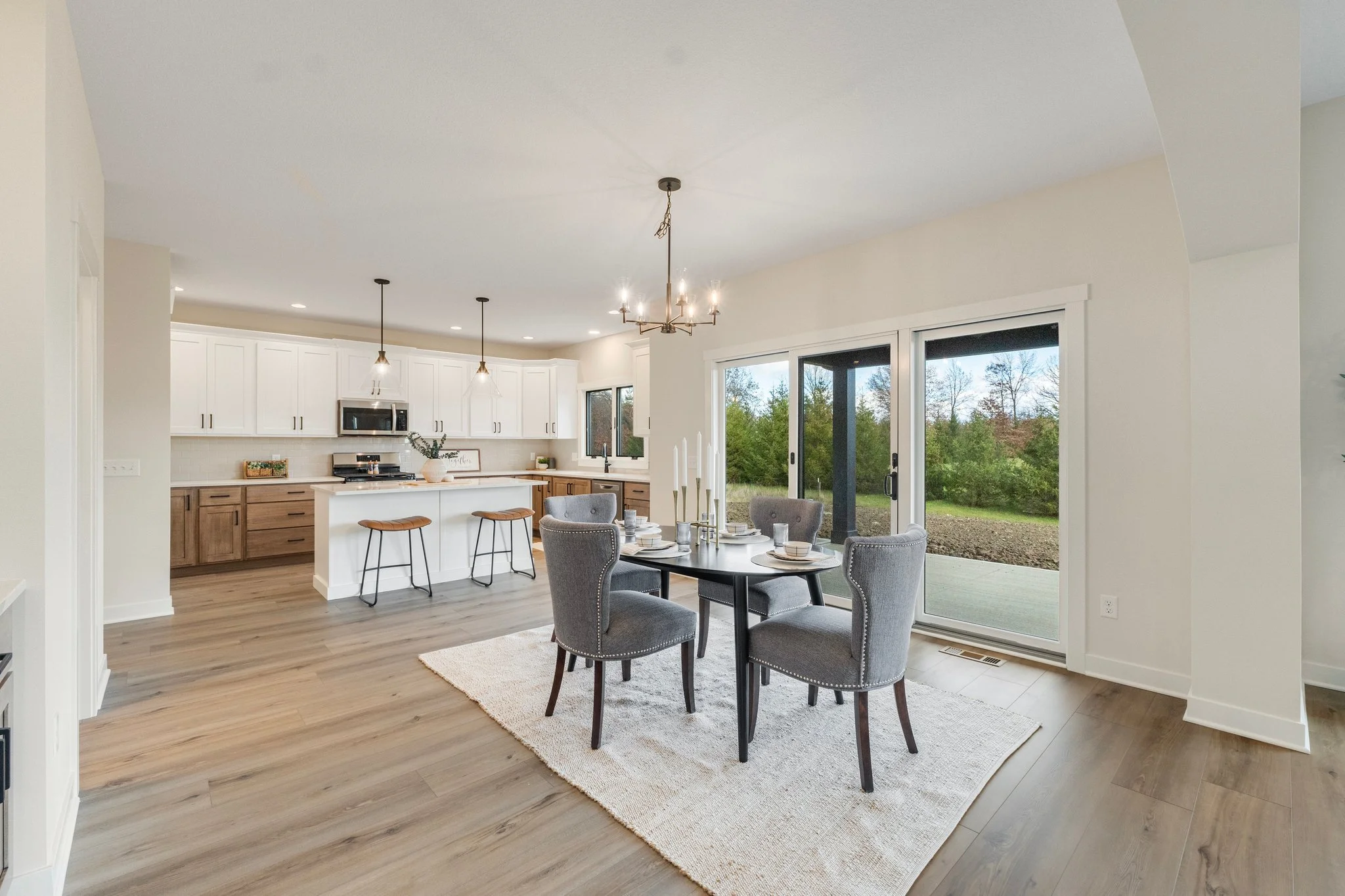 Open-concept kitchen and dining area with sliding glass door leading to outdoors, featuring a black dining table with four gray upholstered chairs, a white kitchen with island, pendant and chandelier lighting, and wood flooring.