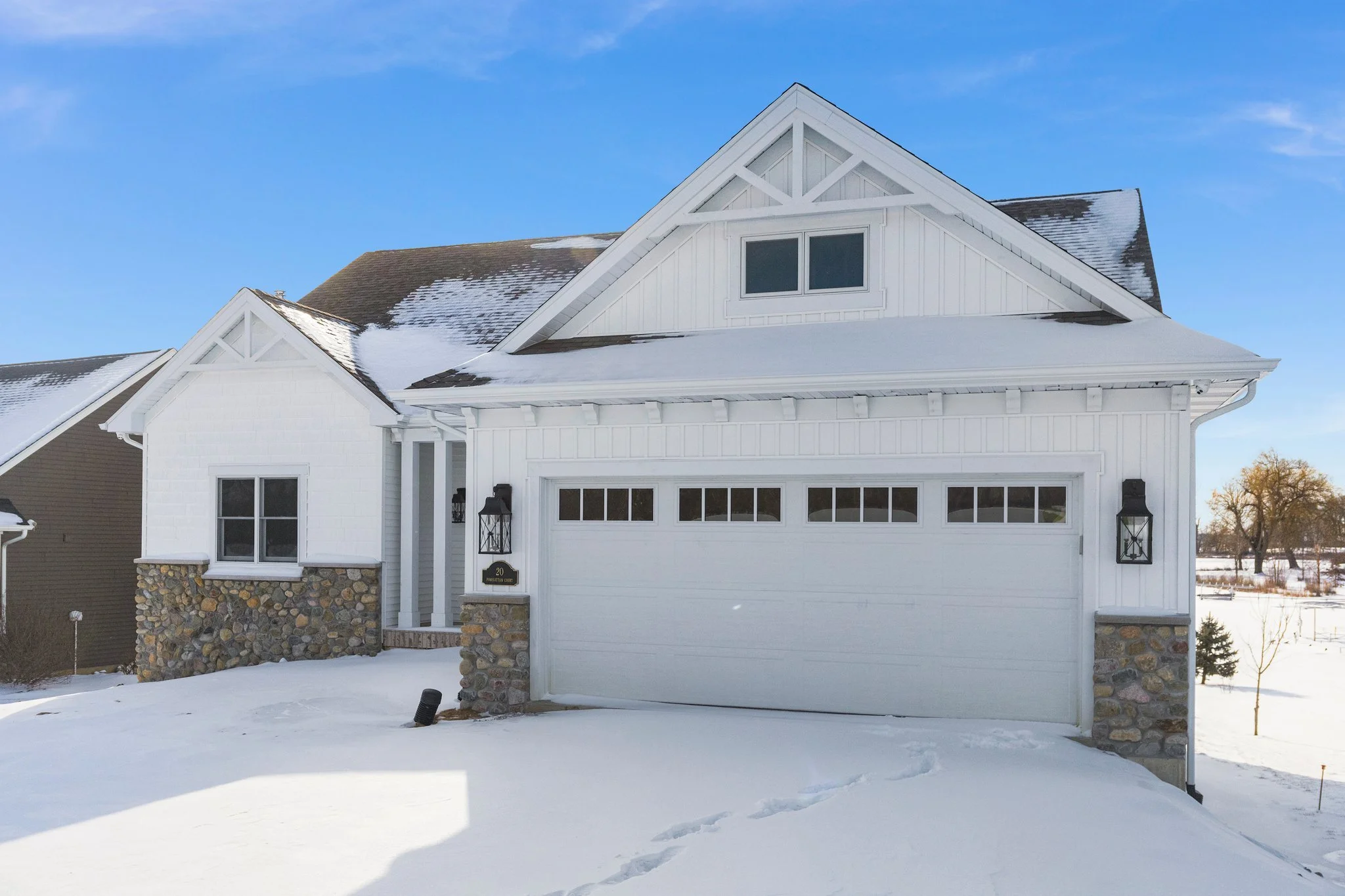 White house with stone accents on the lower walls, a garage door, and snow on the roof and ground, under a blue sky.