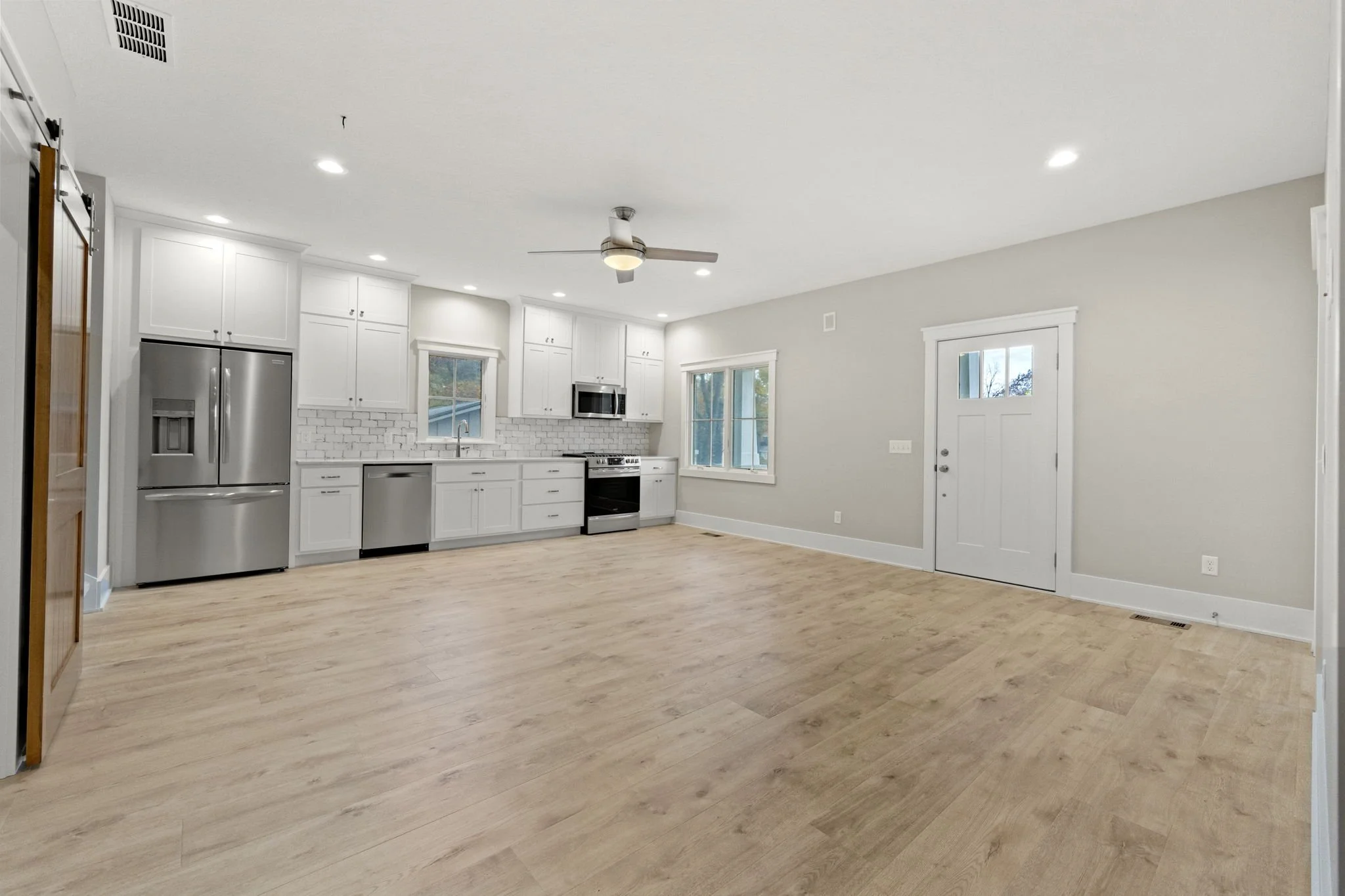 Empty kitchen and living area with white cabinets, stainless steel appliances, light wood flooring, and a ceiling fan.
