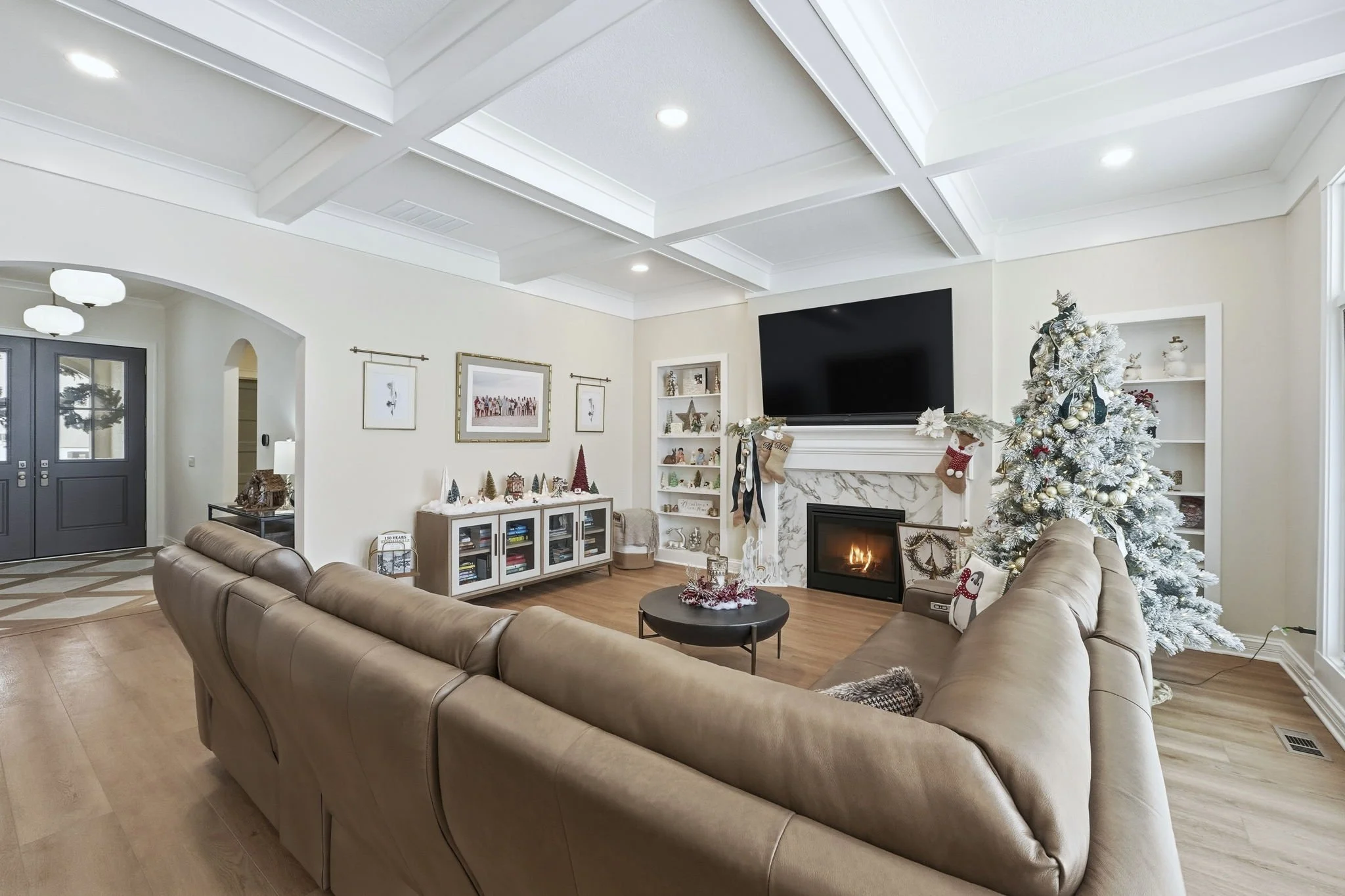 Living room decorated for Christmas with white Christmas tree, stockings, and ornaments, beige sectional sofa, fireplace, mounted television, and various holiday decorations.