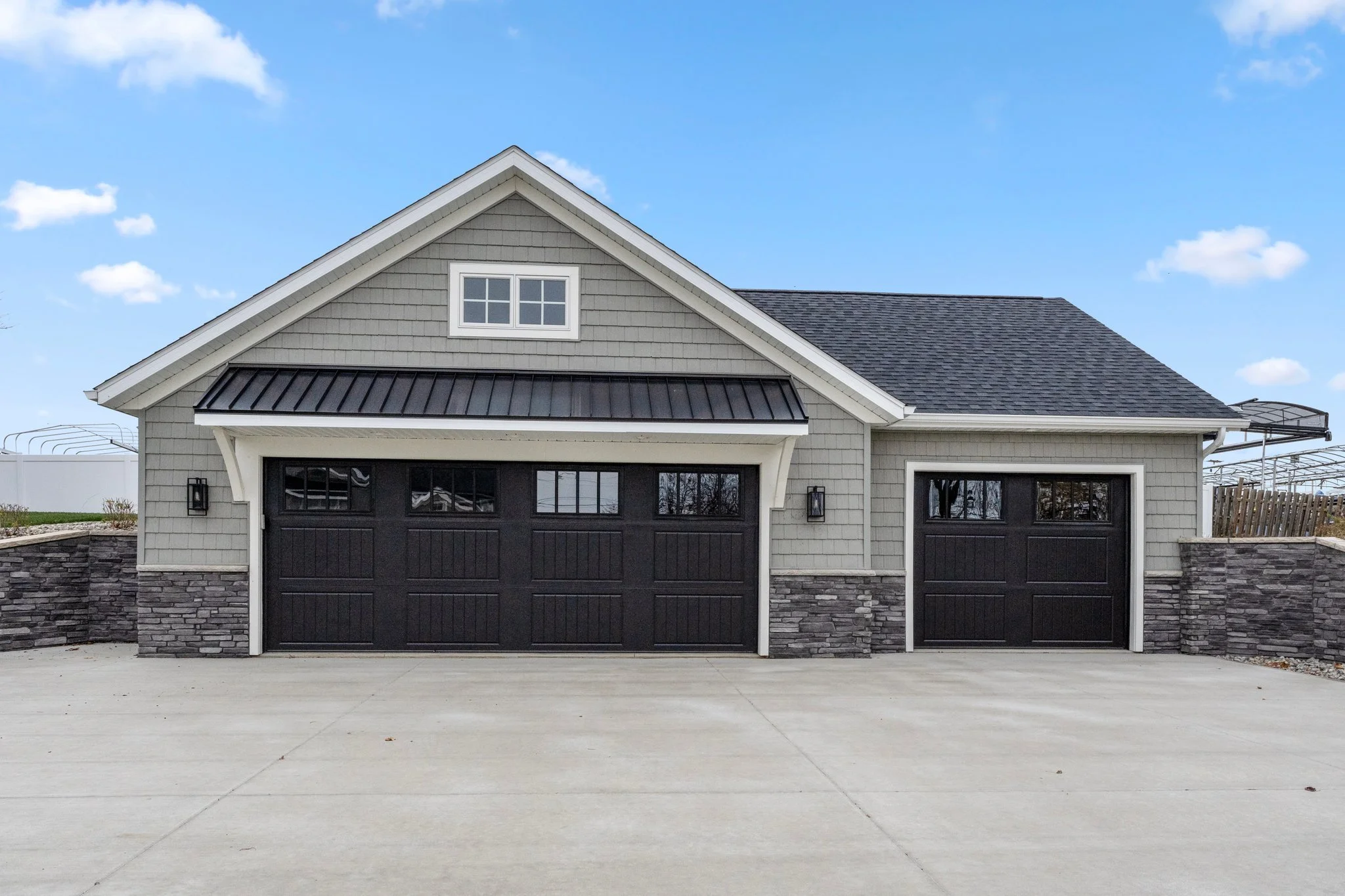 A modern garage with two black doors, grey siding, stone accents, and a gabled roof under a blue sky with clouds.