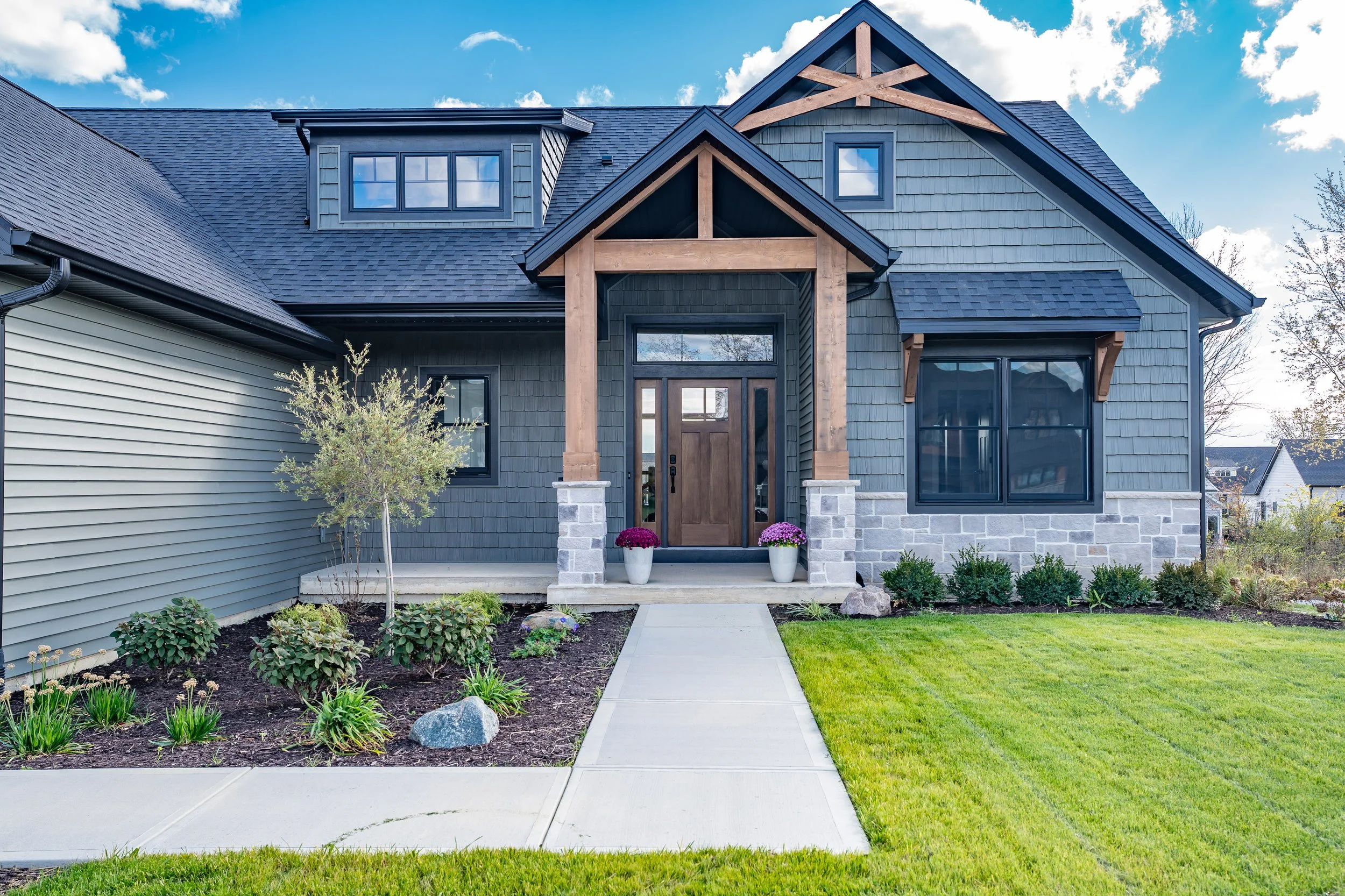 Front view of a modern house with a porch, wooden beams, stone foundation, and black window frames. The yard has green grass, a concrete pathway, and landscaped plants with flowers.