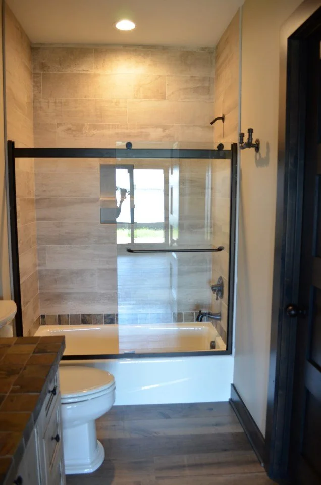 Bathroom with bathtub, glass shower door, toilet, wooden floor, and beige tiled walls.