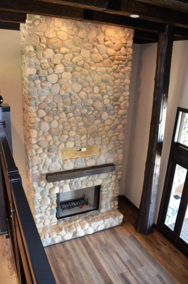 A tall stone fireplace with a wood mantel in a cozy living room, with hardwood flooring and large windows.
