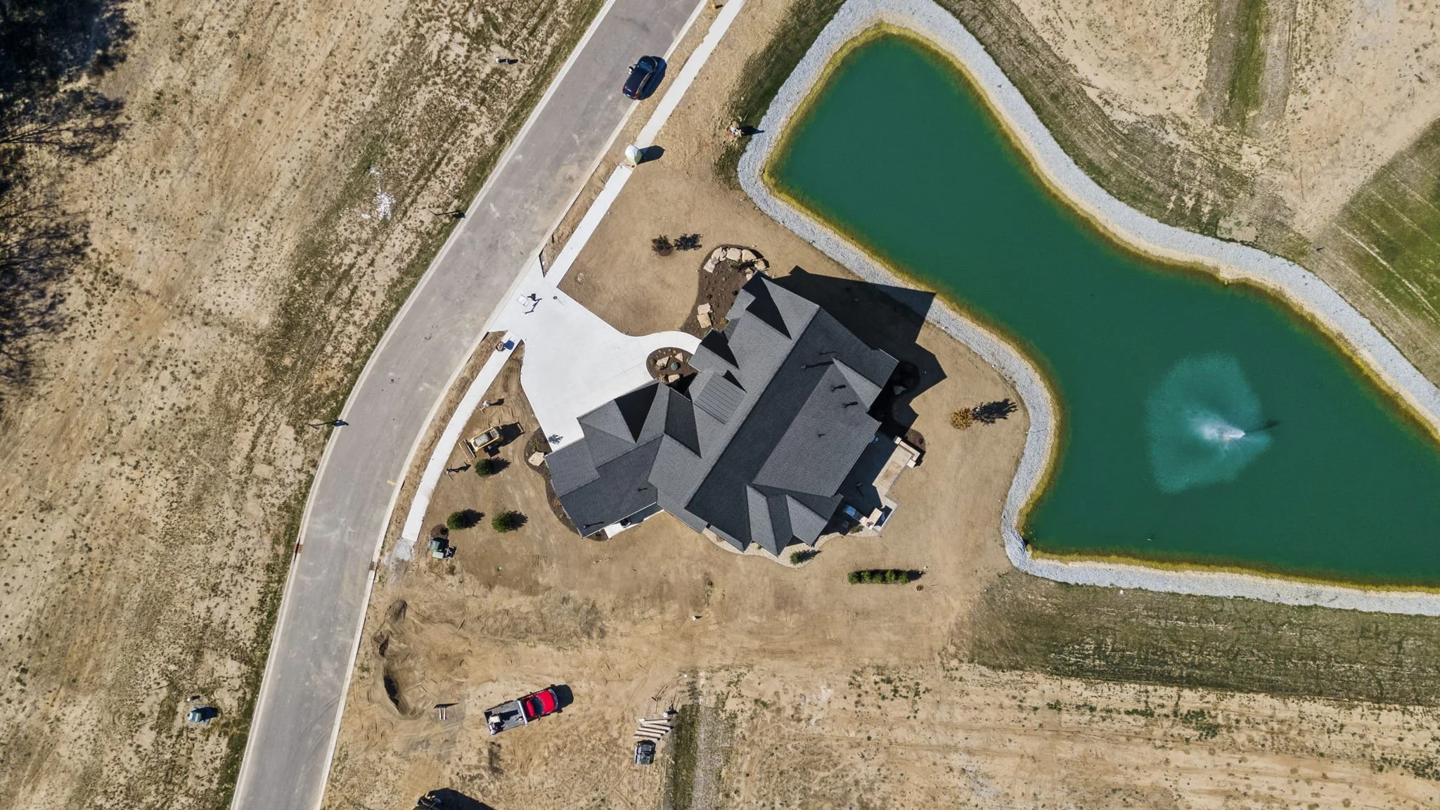 Aerial view of a house with a dark roof next to a pond with a fountain, surrounded by a lawn and a road with parked cars.