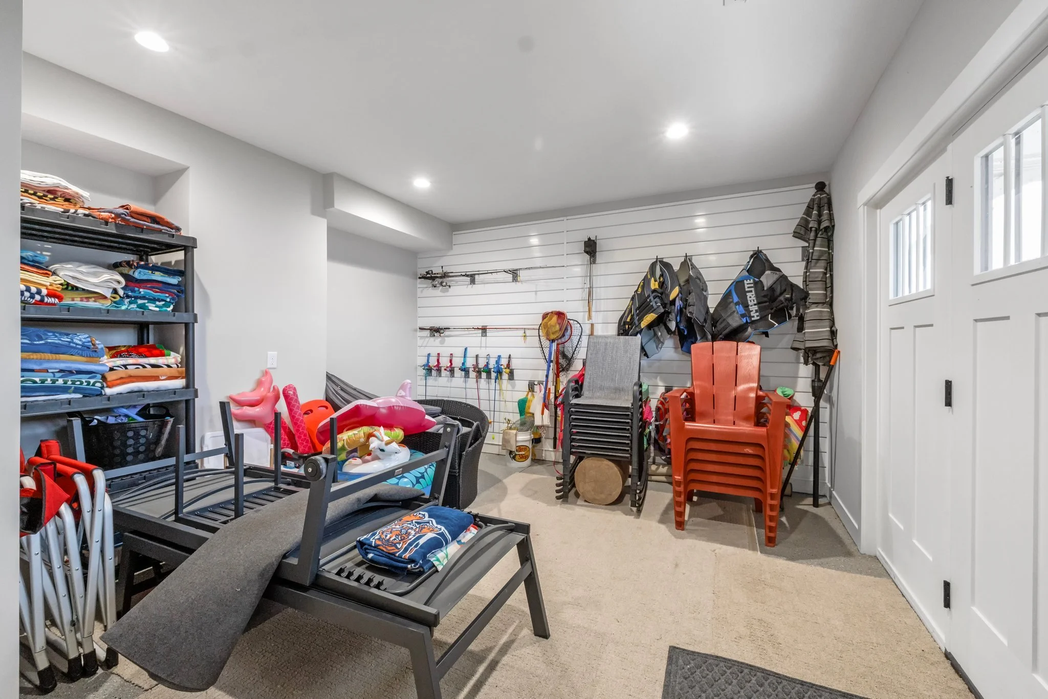 Garage with shelves of folded clothes, chairs stacked, various toys including inflatable swan, and stored outdoor gear like fishing rods, life jackets, and umbrellas.
