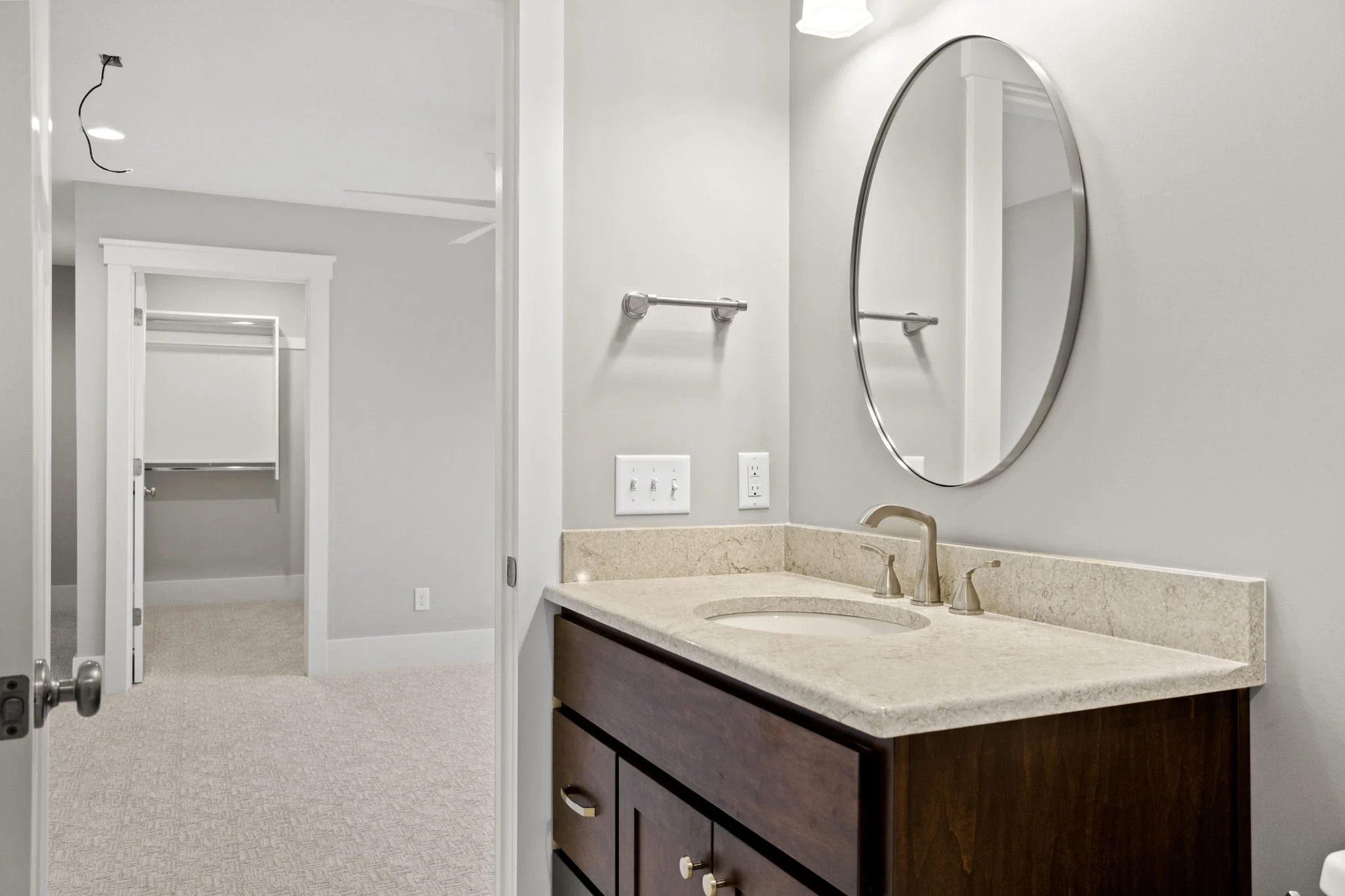 Bathroom vanity with a beige countertop, dark wood cabinet, oval mirror, and a faucet, with a towel bar and electrical outlets on the wall. A white door leads to a closet with white shelves in the background.